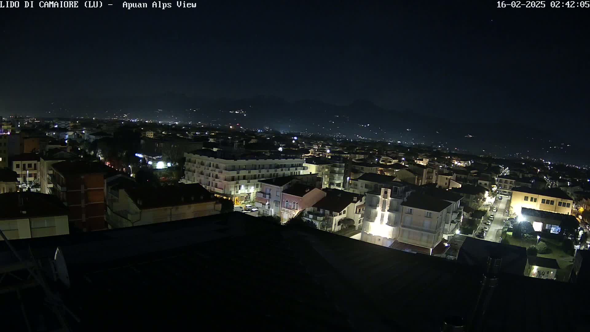 A nighttime aerial view shows a town illuminated by streetlights, with mountains visible in the distance under a clear, dark sky.