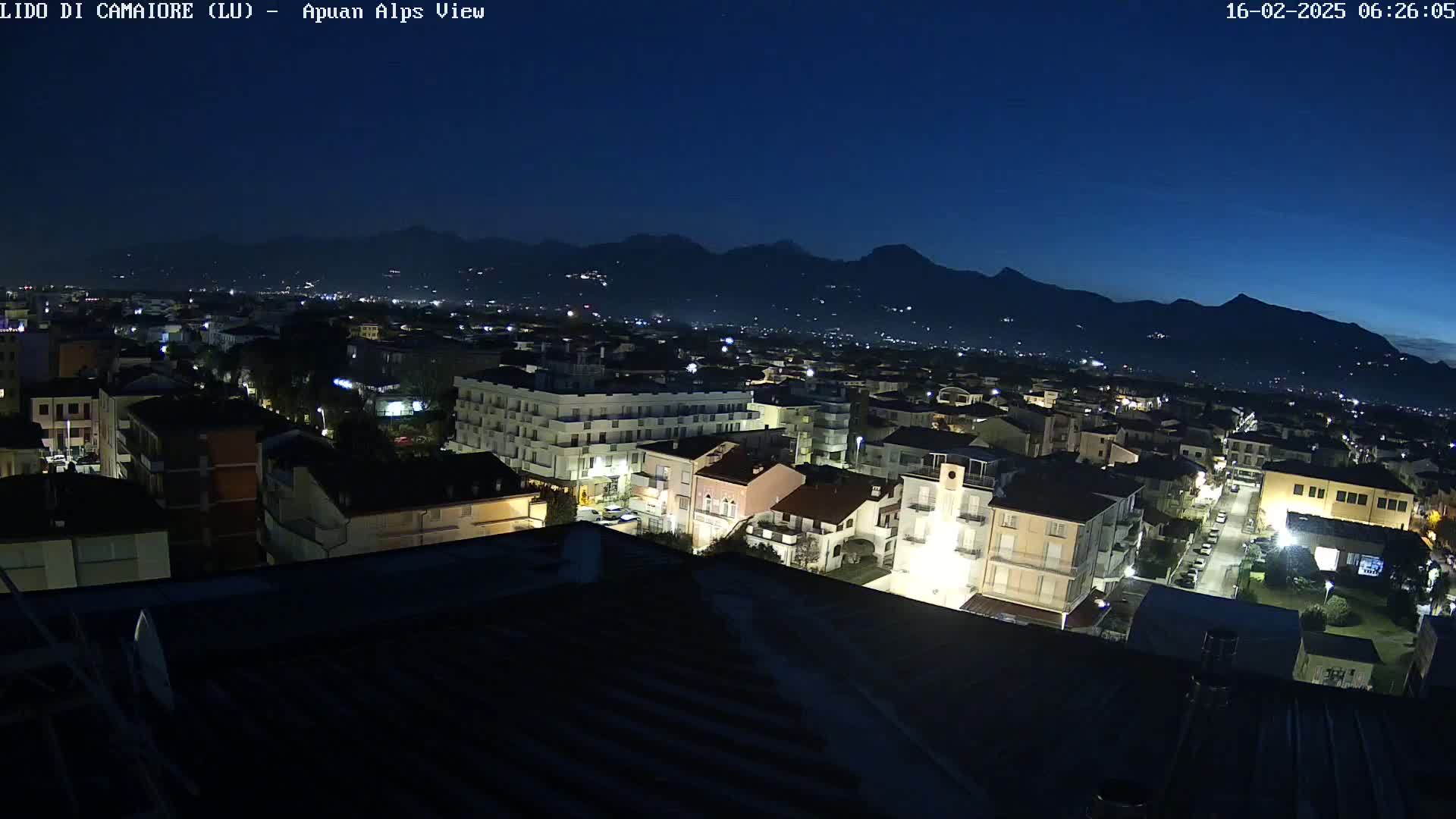 A nighttime, high-angle view of a town illuminated by streetlights, nestled against a dark mountain range under a clear, dark blue sky.