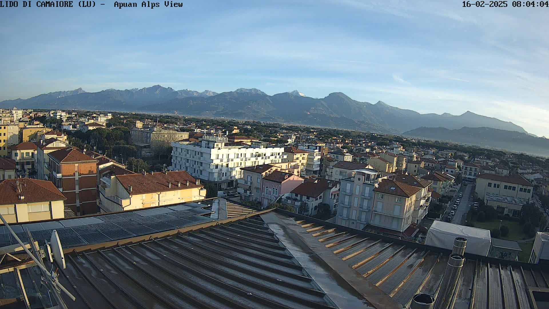 A rooftop view shows a town nestled in a valley with snow-capped mountains in the background under a clear, sunny sky.