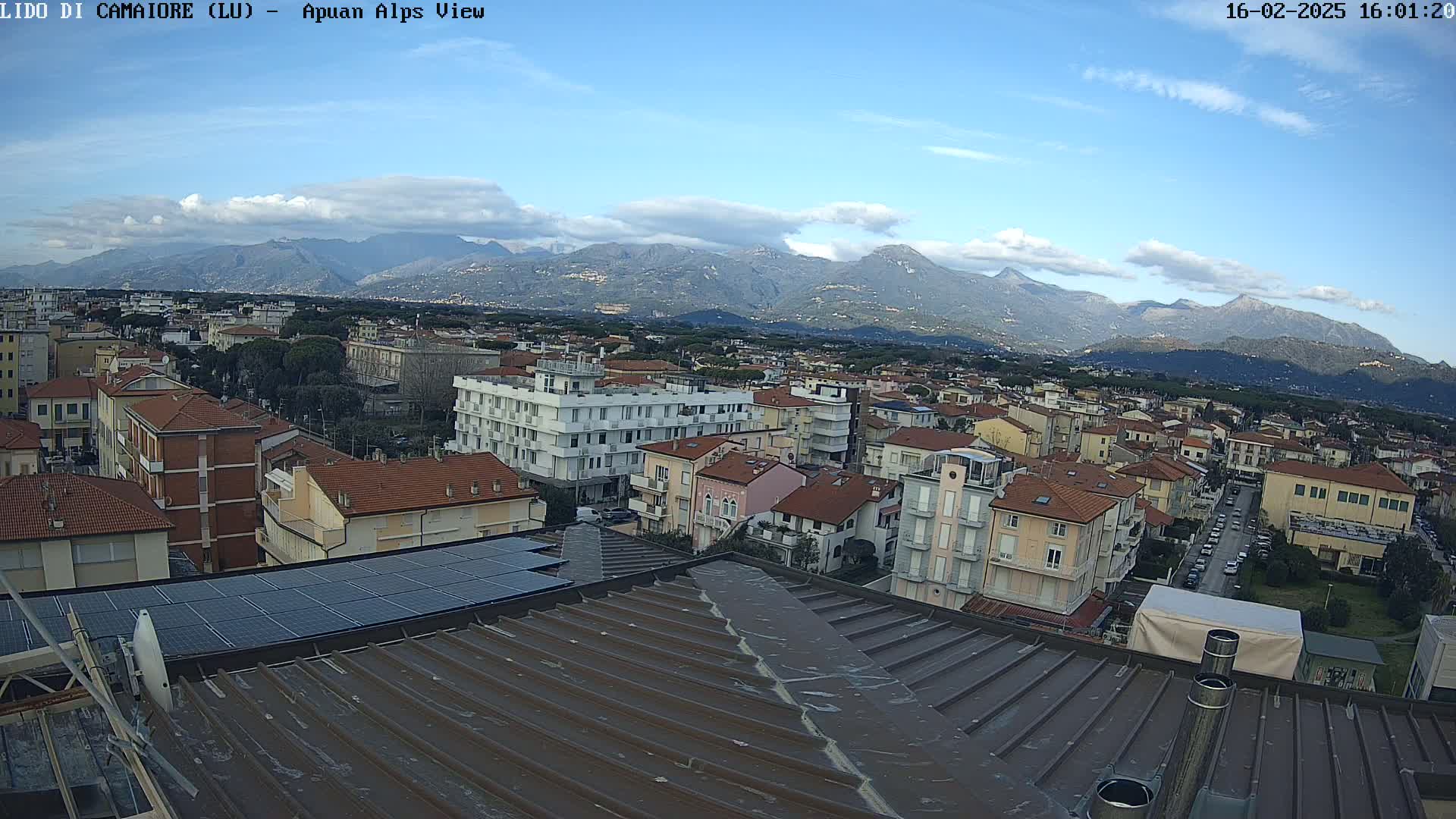 A mostly sunny day reveals a coastal town nestled below a mountain range, viewed from a rooftop with solar panels.