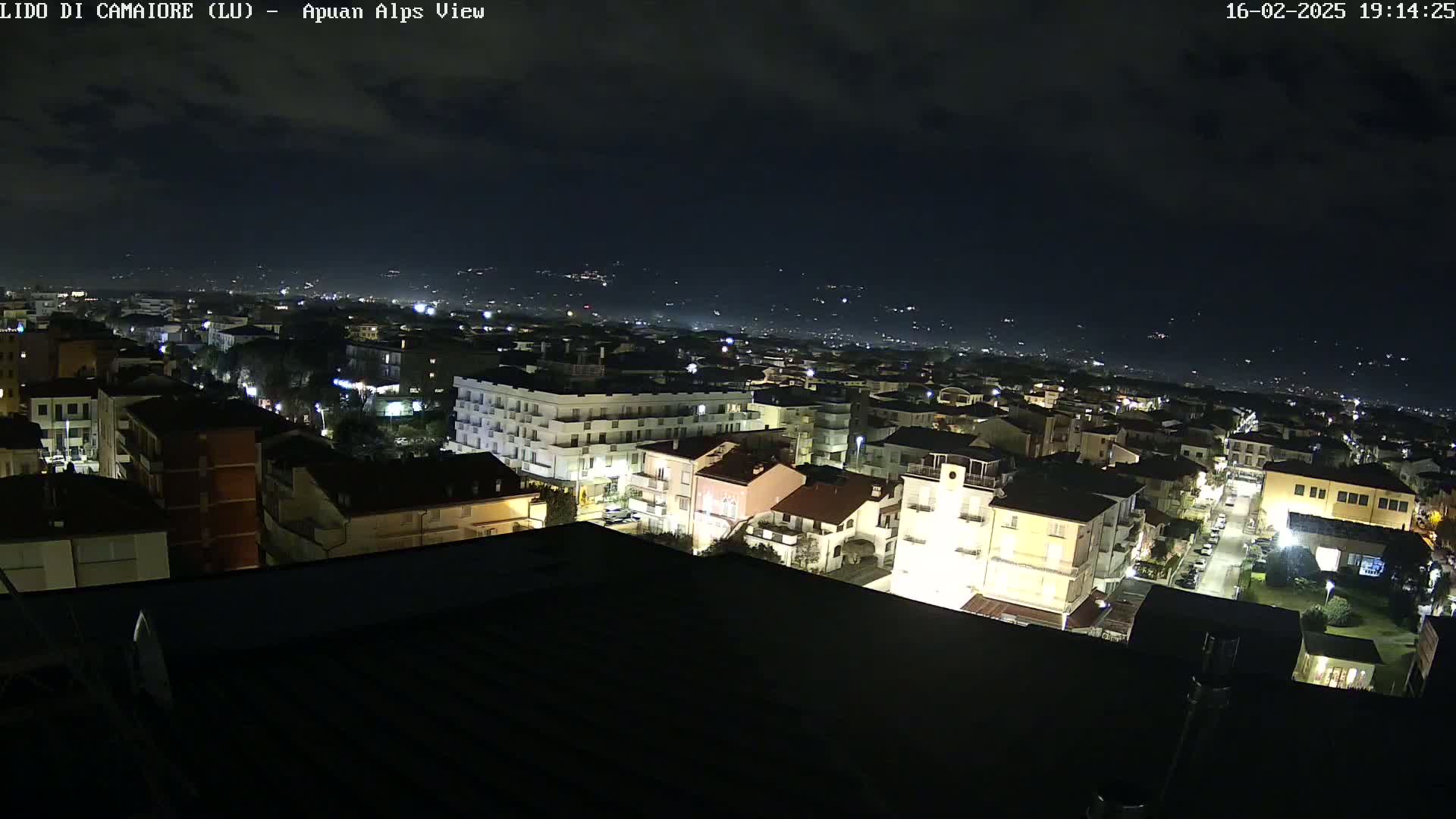 A nighttime aerial view of a town shows buildings illuminated, with distant lights in the background under a mostly clear sky.