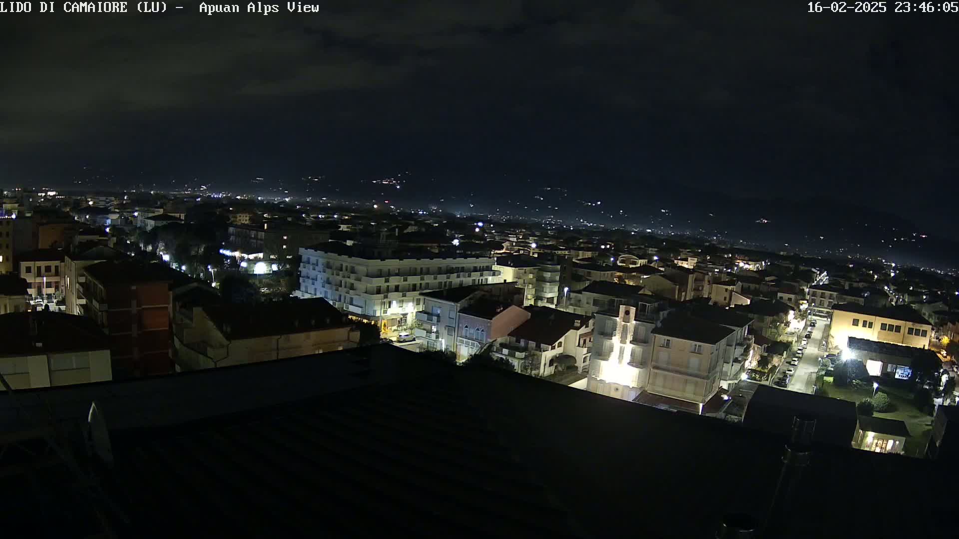 A nighttime, aerial view of a town illuminated by streetlights, with dark mountains visible in the distance under a mostly clear sky.