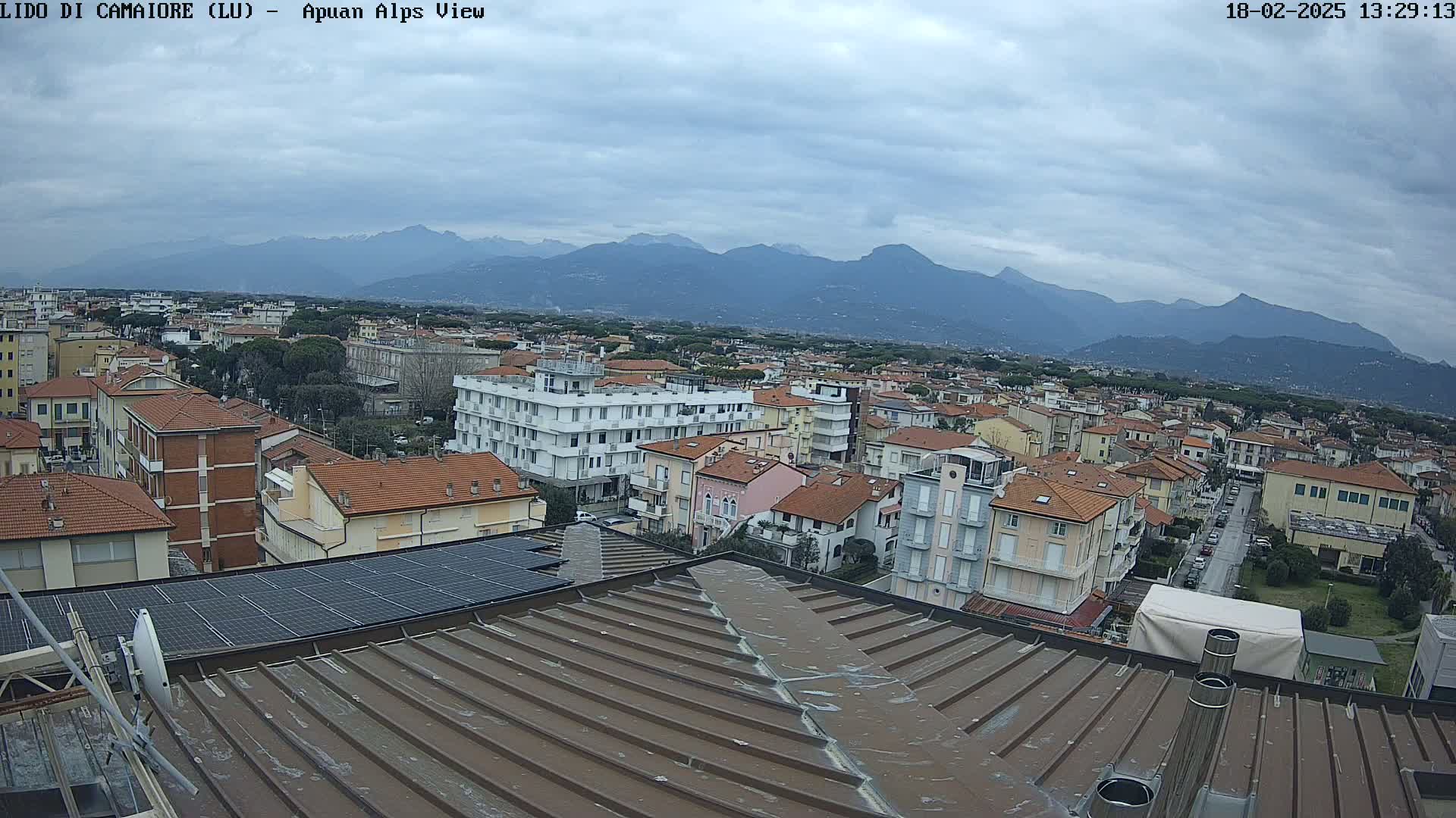 A view from a rooftop shows a town of low-rise buildings under a cloudy sky, with mountains visible in the distance.