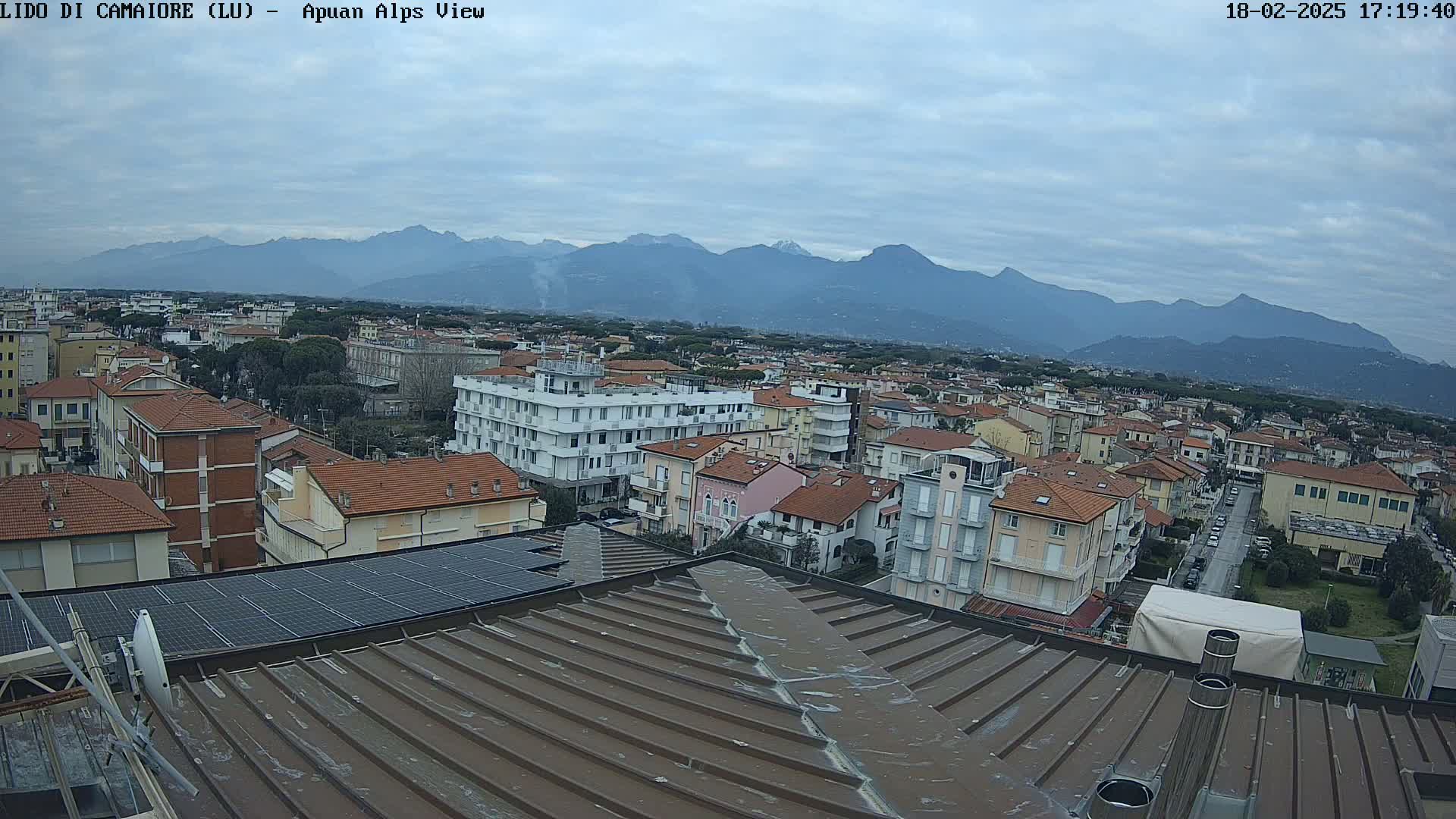 A view of a town with red-tiled roofs, under an overcast sky, with mountains in the background.