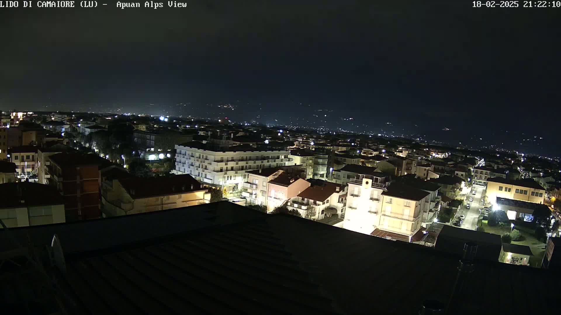 A nighttime, high-angle view of a town's buildings illuminated by streetlights, with a dark sky and distant lights visible on the horizon.