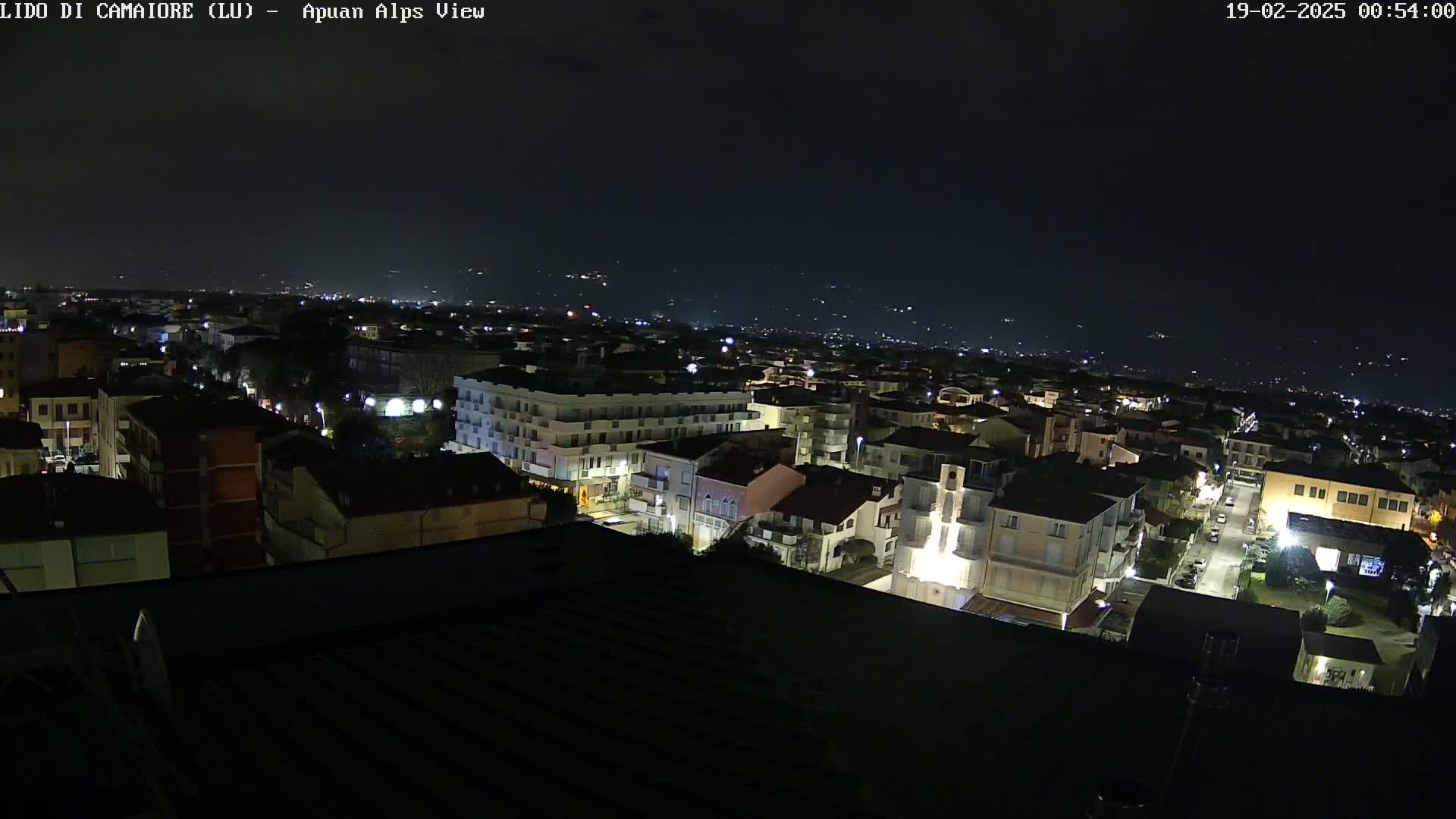 A nighttime, aerial view of a town with illuminated buildings and streets, extending to a distant, dimly lit landscape under a dark sky.
