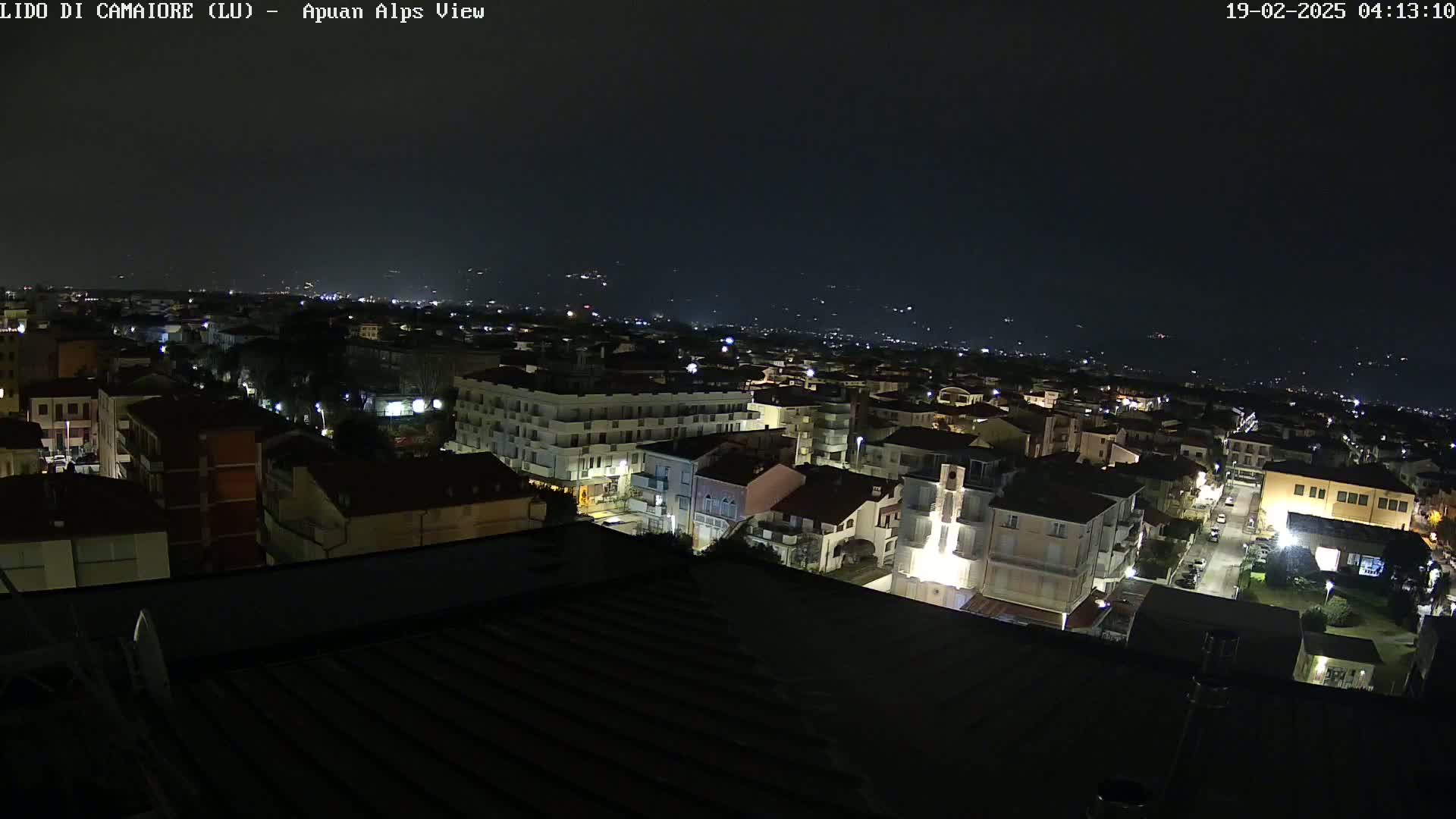 A nighttime aerial view of a town shows numerous buildings illuminated by streetlights, with a dark sky and distant lights suggesting a clear night.