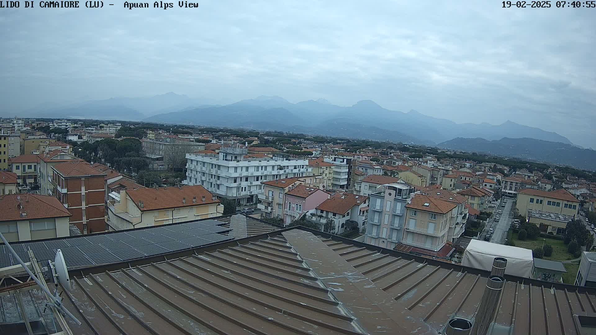 A view from a rooftop shows a town with numerous buildings under a cloudy sky, with distant mountains visible in the background.
