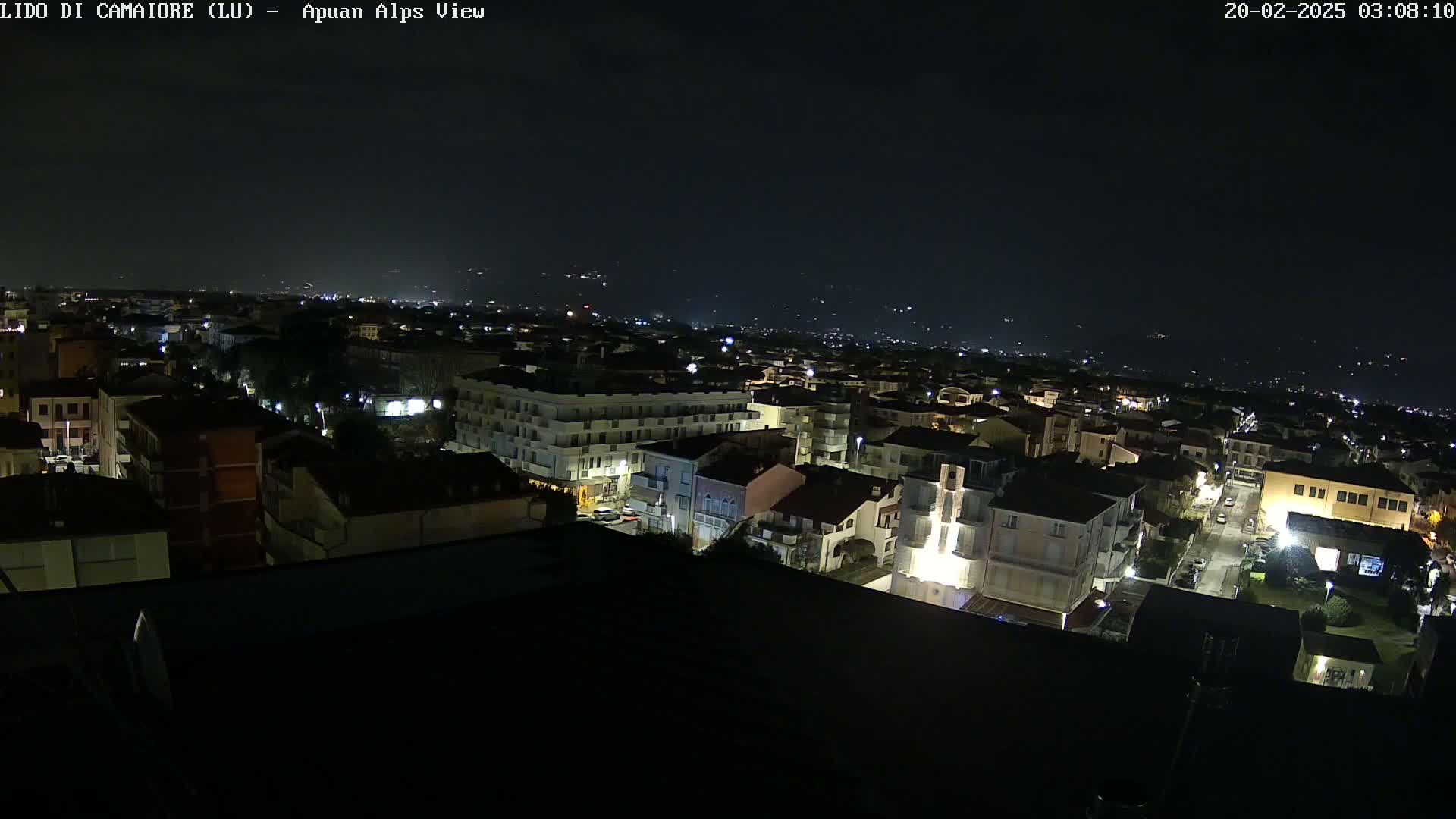 A nighttime aerial view of a town shows many buildings with sparsely lit streets, and distant hills faintly visible under a dark sky.