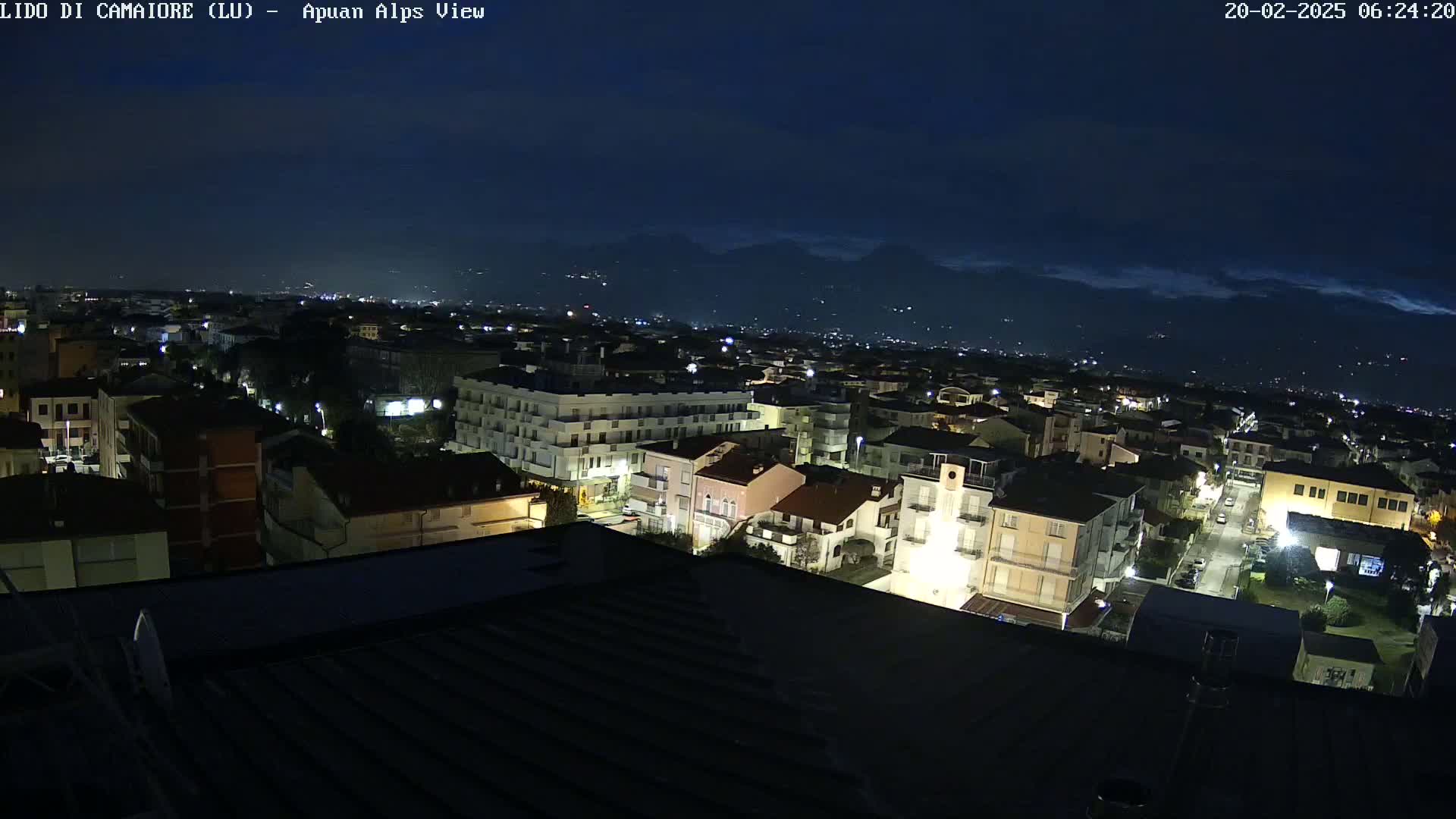 A nighttime view of a town illuminated by streetlights, with mountains visible in the distance under a dark, cloudy sky.