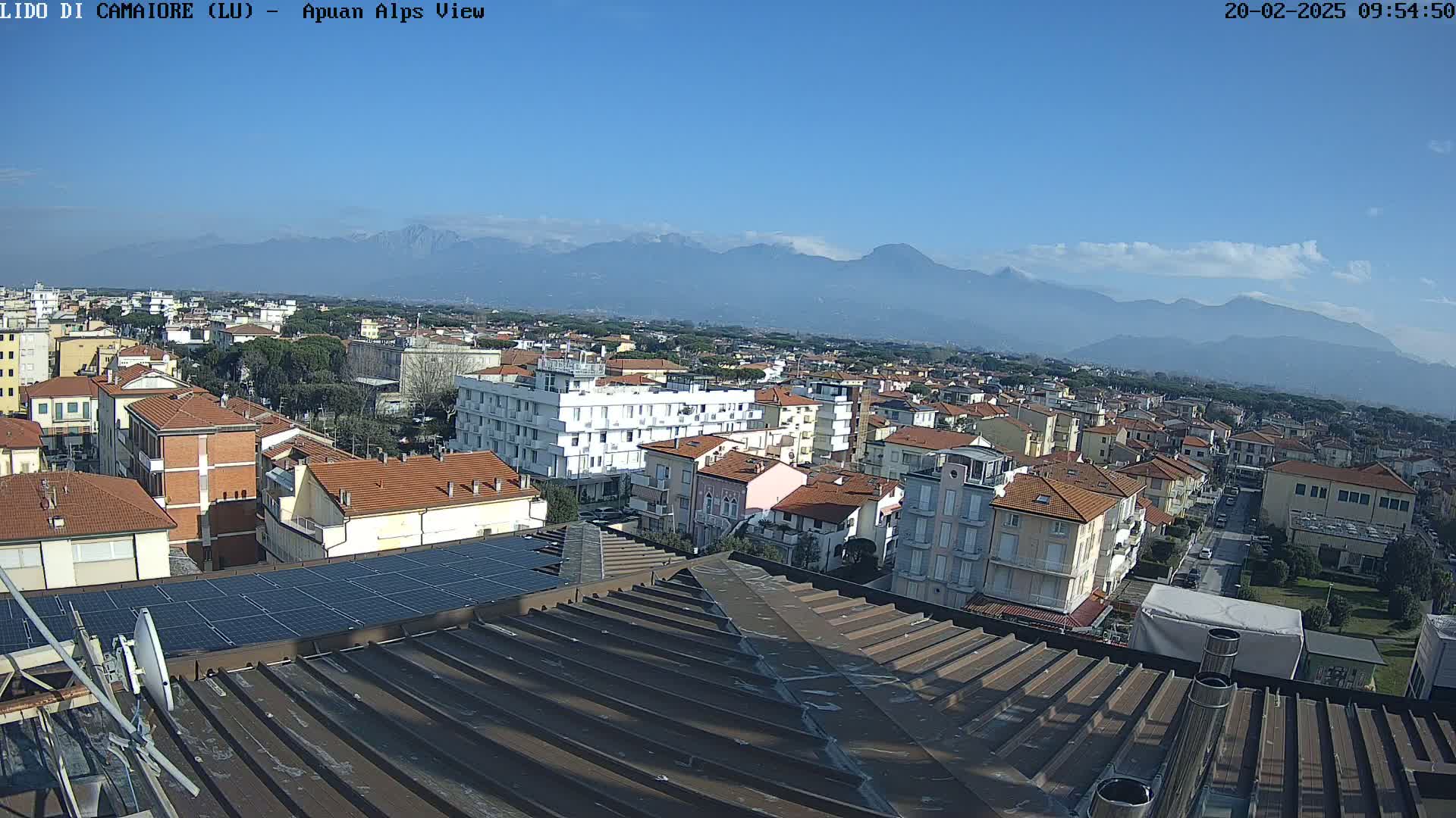 A clear, sunny day reveals a cityscape of low-rise buildings and a distant mountain range under a mostly clear blue sky.