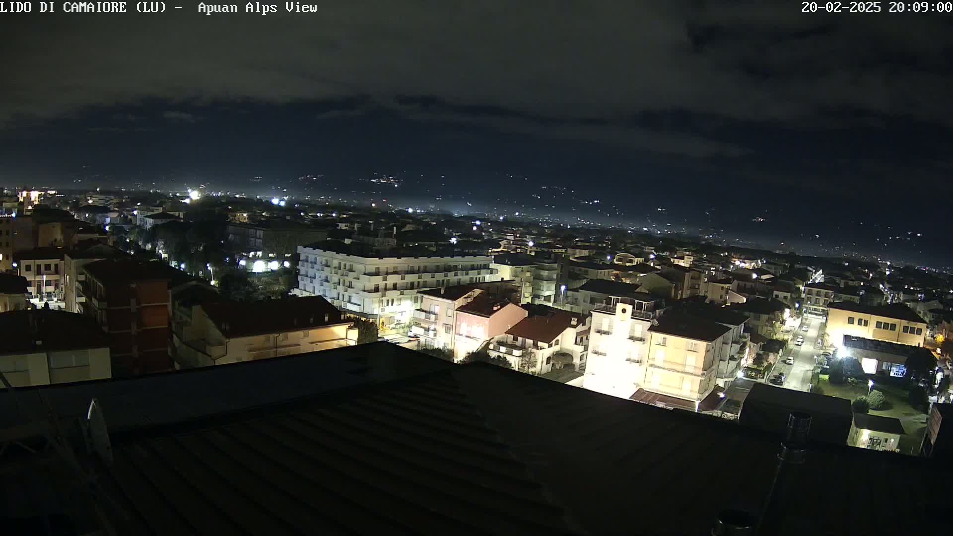 A nighttime, high-angle view of a town illuminated by streetlights, with a hazy sky and distant lights indicating other settlements in the background.