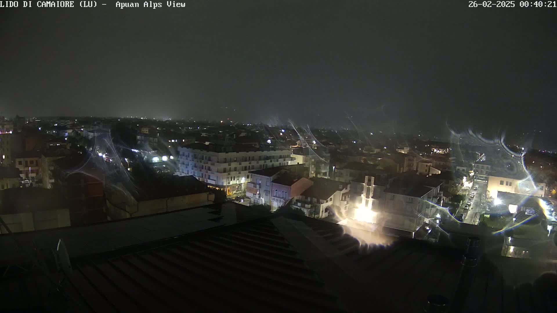 A nighttime view of a city illuminated by streetlights, with streaks of rain or snow visible in the dark sky.