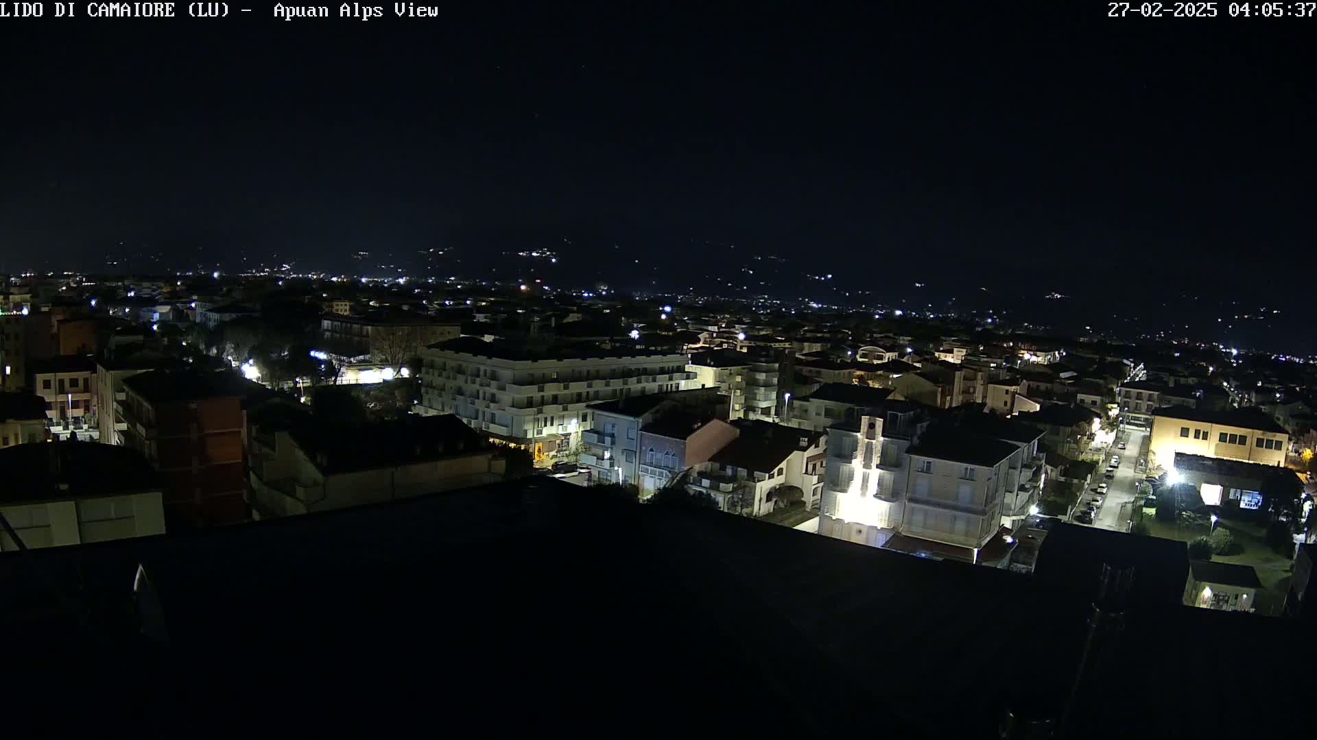 A nighttime, high-angle view of a town's illuminated buildings and streets, with distant hills faintly visible under a clear, dark sky.