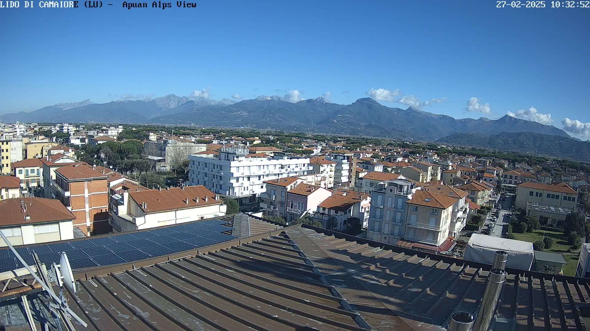 A mostly sunny day reveals a cityscape nestled against a backdrop of distant mountains, viewed from a rooftop with solar panels.