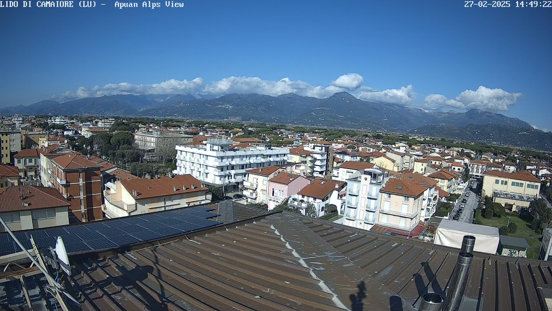 A rooftop view of a town shows mostly low-rise buildings under a mostly sunny sky with some clouds, and mountains in the background.