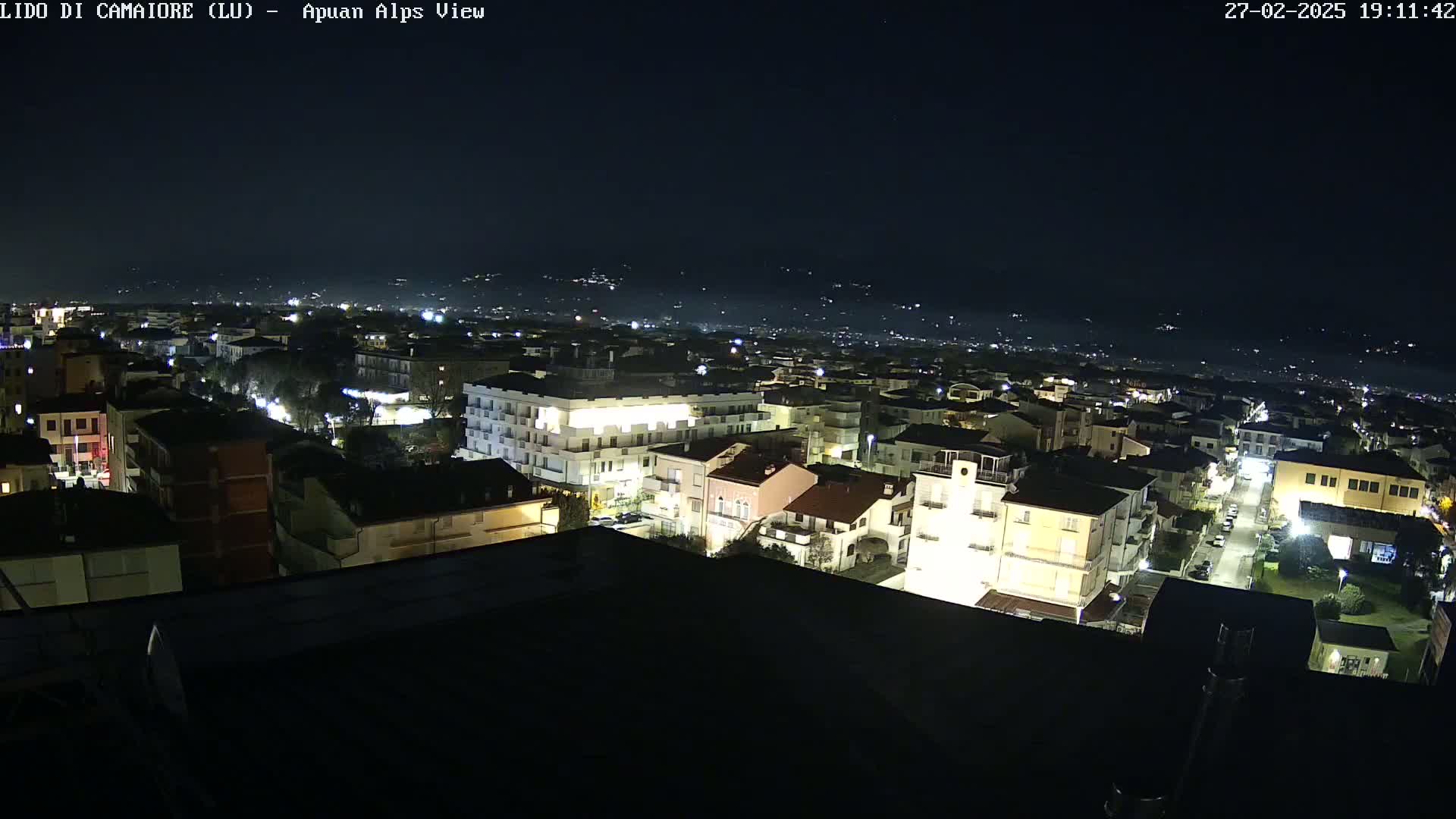 A nighttime aerial view of a town illuminated by streetlights and building lights, with a dark, clear sky and distant hills faintly visible in the background.