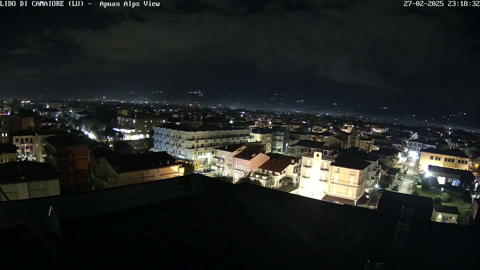 A nighttime aerial view of a town, illuminated by streetlights, with a dark, cloudy sky and distant mountains visible in the background.