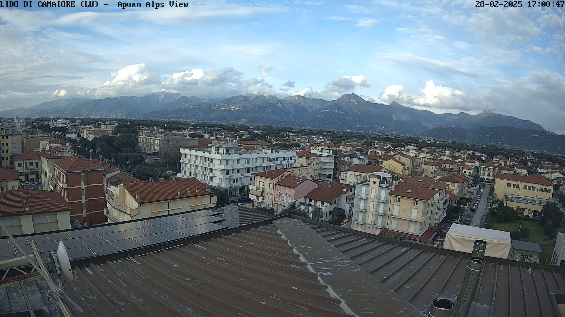 A partly cloudy day reveals a cityscape nestled beneath a mountain range, viewed from a rooftop with solar panels.