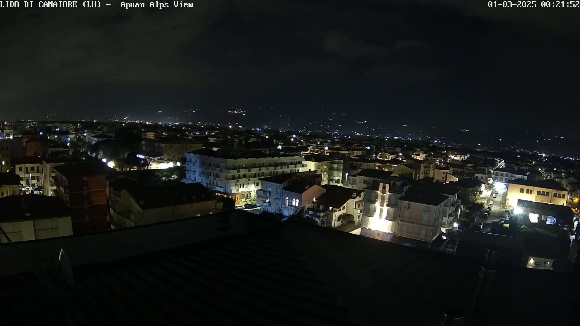 A nighttime aerial view shows a town illuminated by streetlights, with distant hills visible under a partly cloudy sky.