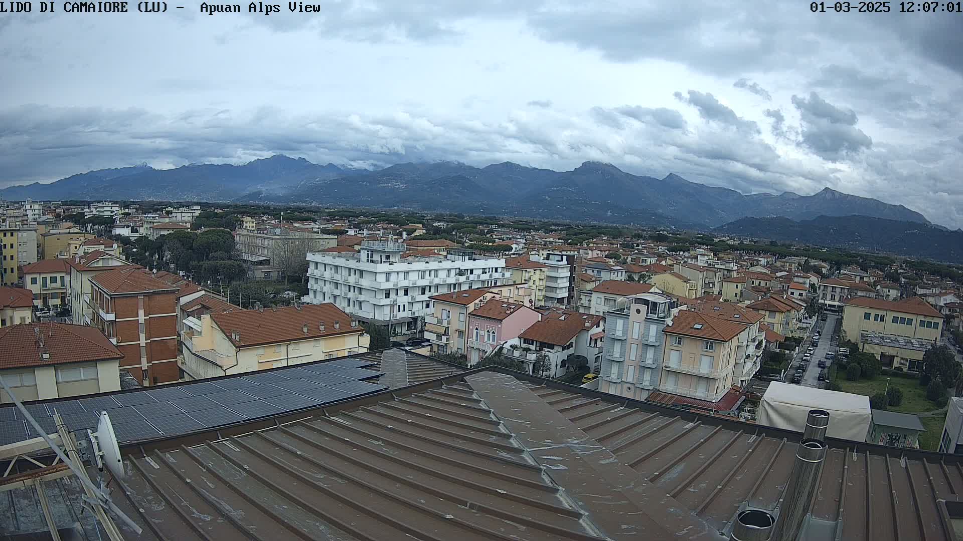 A view from a rooftop shows a town with many low-rise buildings, under a cloudy sky with mountains in the distance.