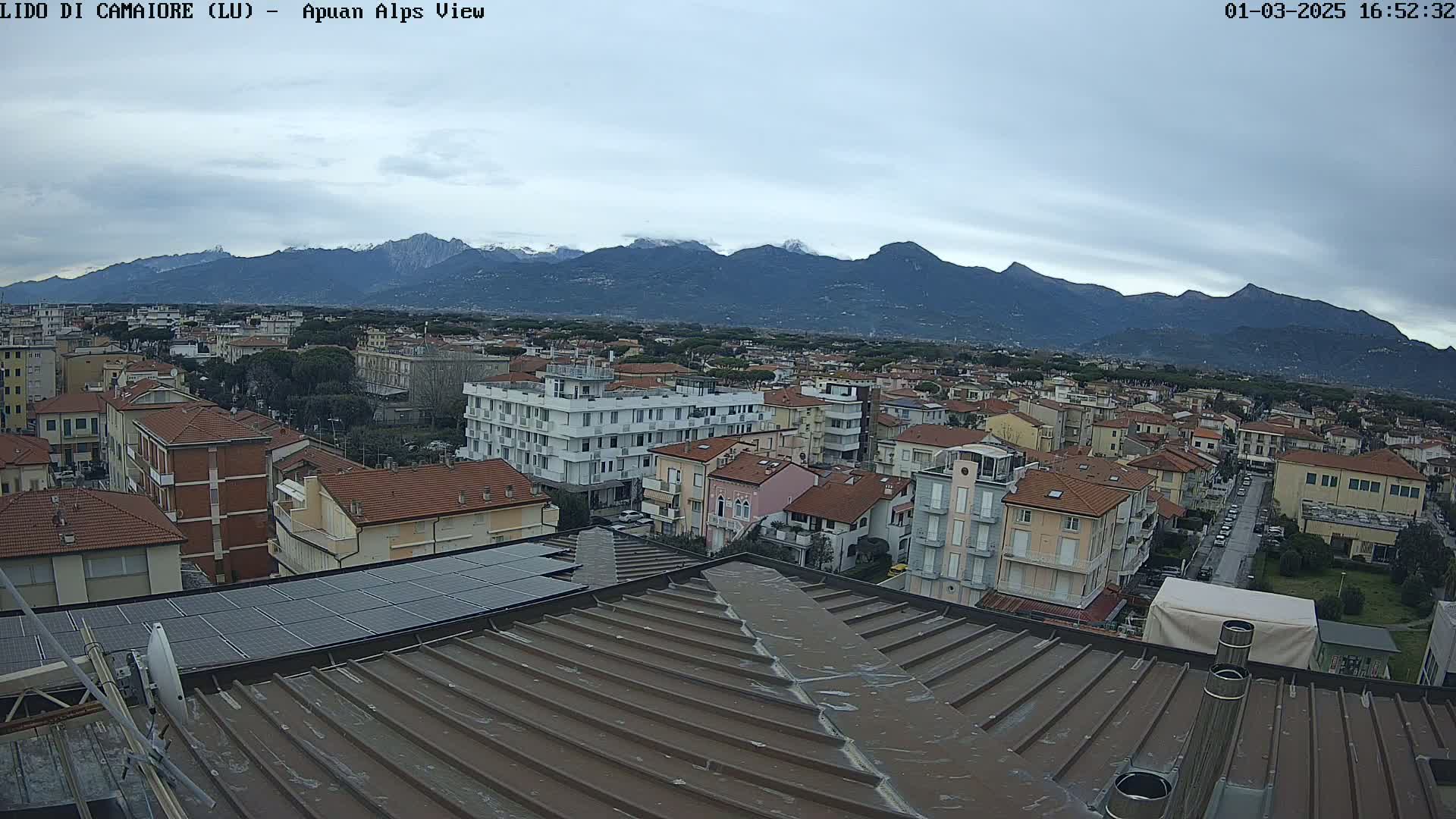 A view from a rooftop shows a town of low-rise buildings stretching toward a mountain range under an overcast sky.