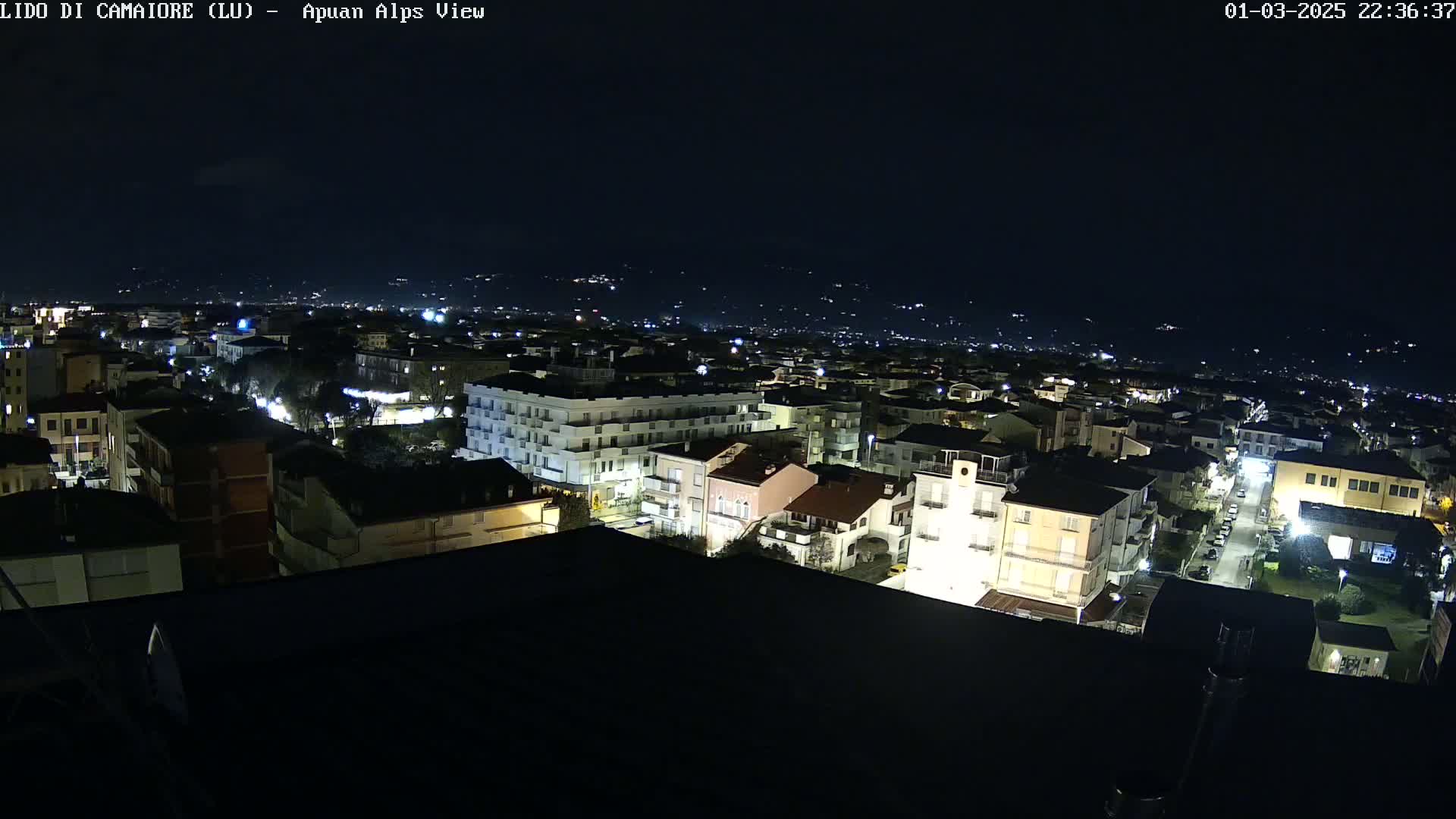 A nighttime, high-angle view of a town's illuminated buildings and streets, with distant, dimly lit hills in the background under a clear, dark sky.