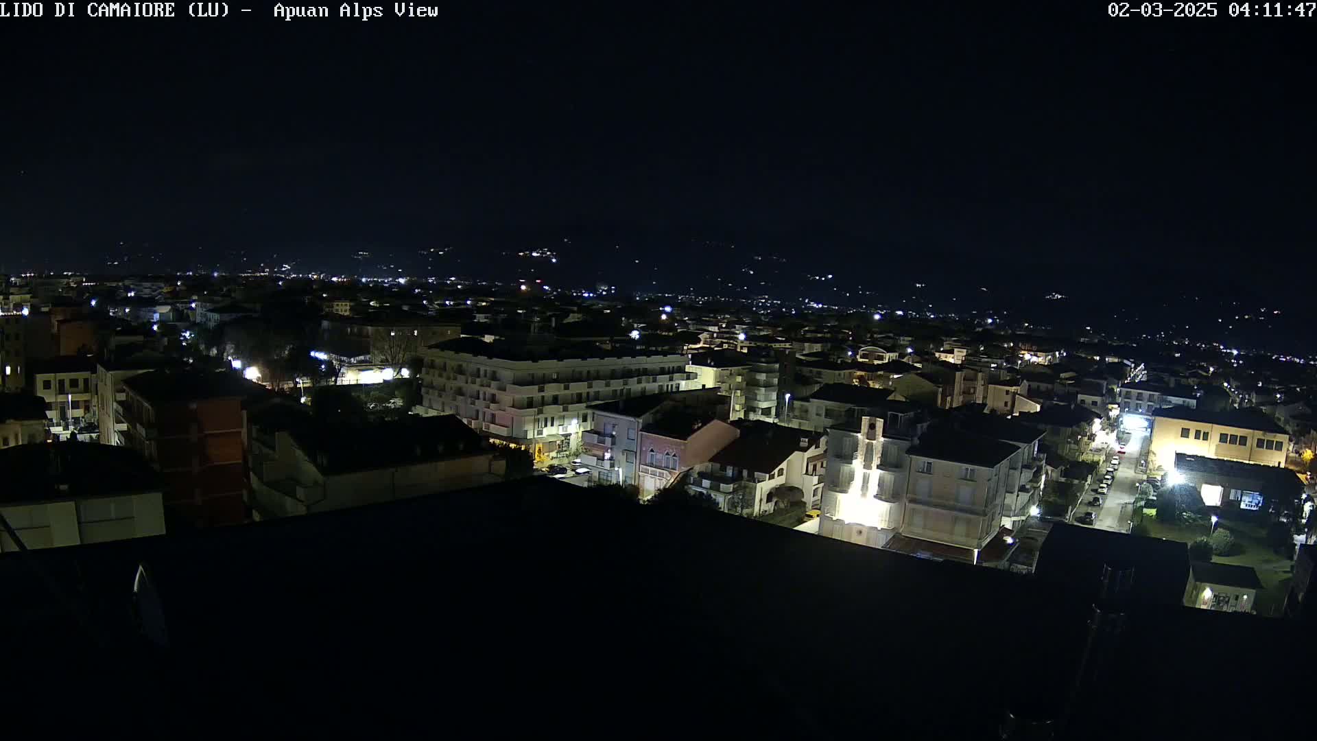 A nighttime aerial view of a town, illuminated by streetlights, with a distant mountain range visible under a clear, dark sky.