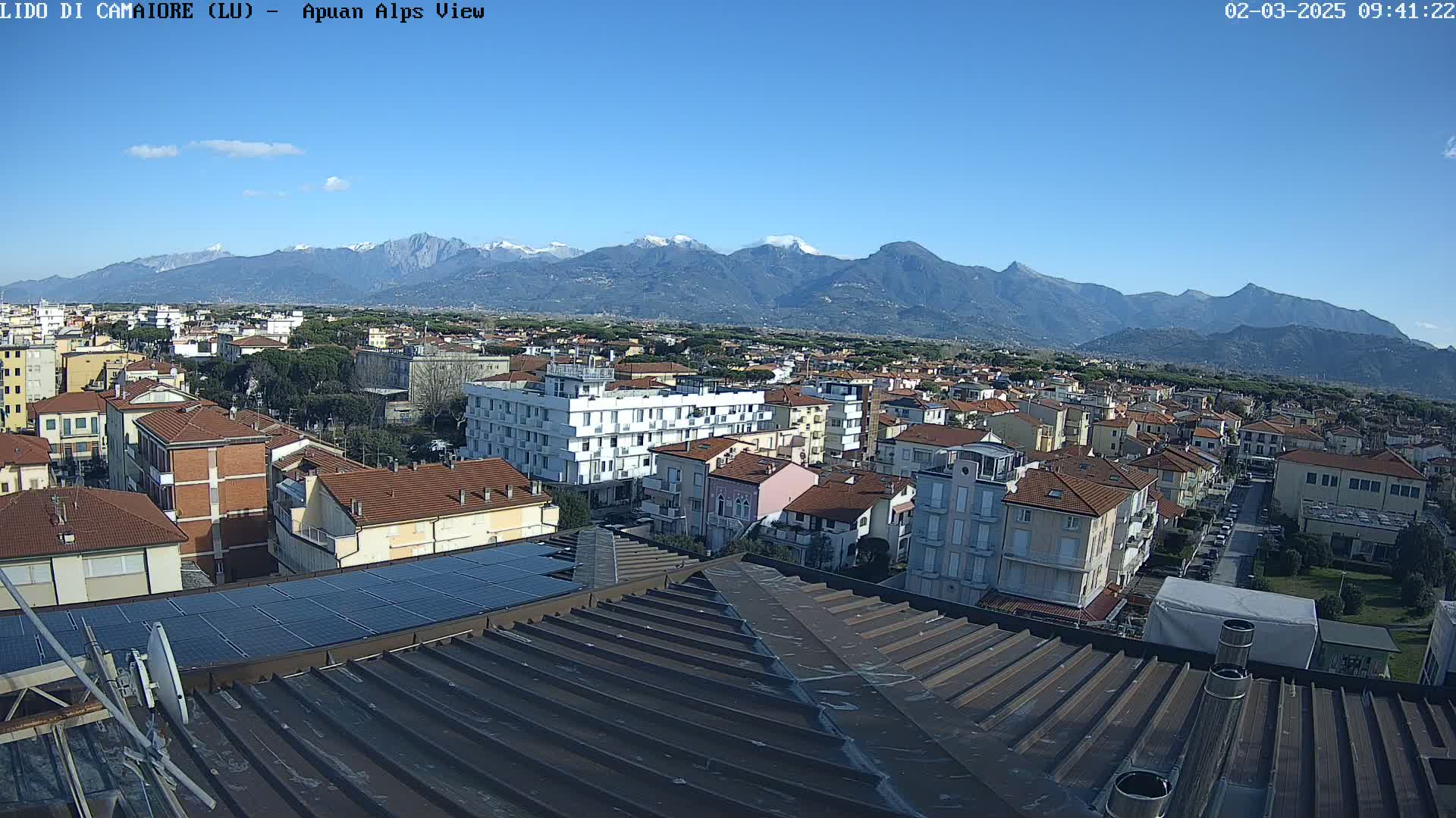 A clear, sunny day reveals a cityscape nestled against a backdrop of snow-capped mountains.