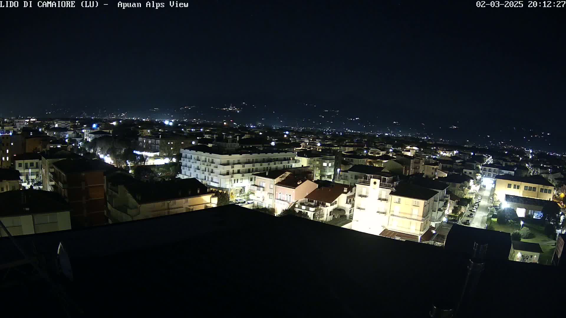 A nighttime, aerial view shows a town's illuminated buildings and streets, with distant hills visible under a clear, dark sky.