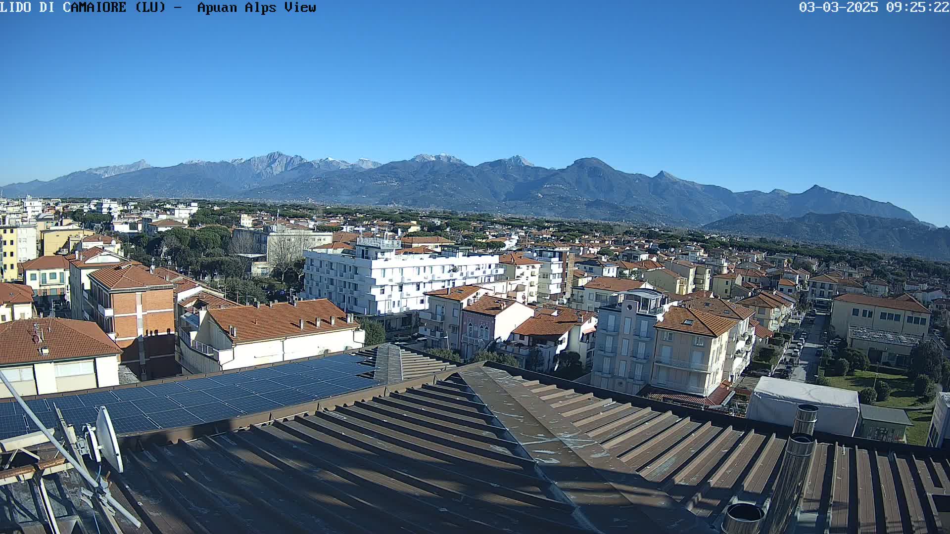 A sunlit cityscape of low-rise buildings stretches towards snow-capped mountains under a clear blue sky.