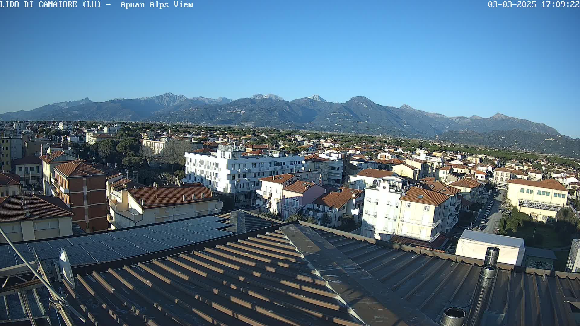A clear, sunny day reveals a panoramic view of a town nestled at the foot of a snow-capped mountain range.