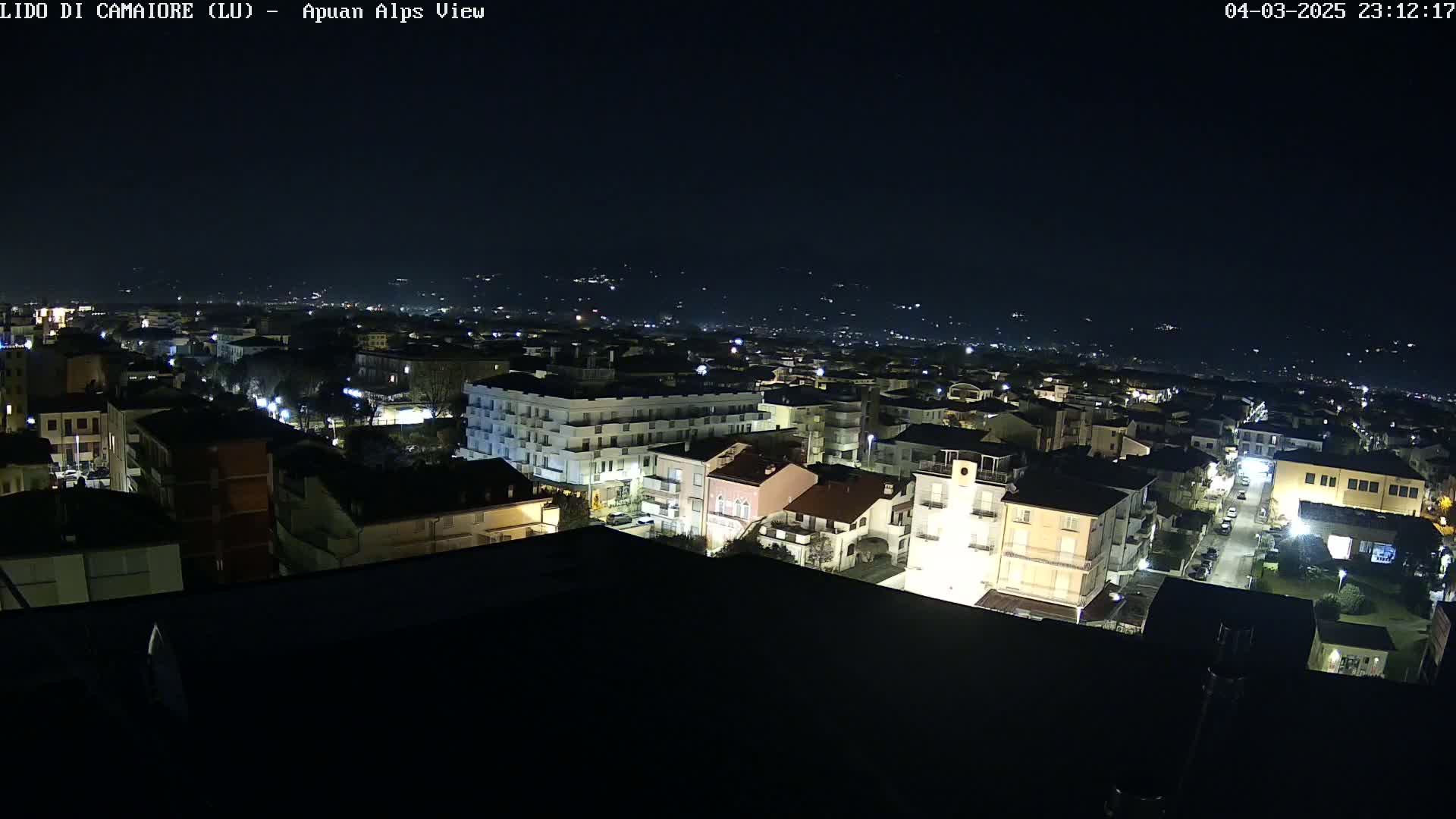 A nighttime aerial view of a city illuminated by streetlights, with a dark sky and distant hills faintly visible in the background.