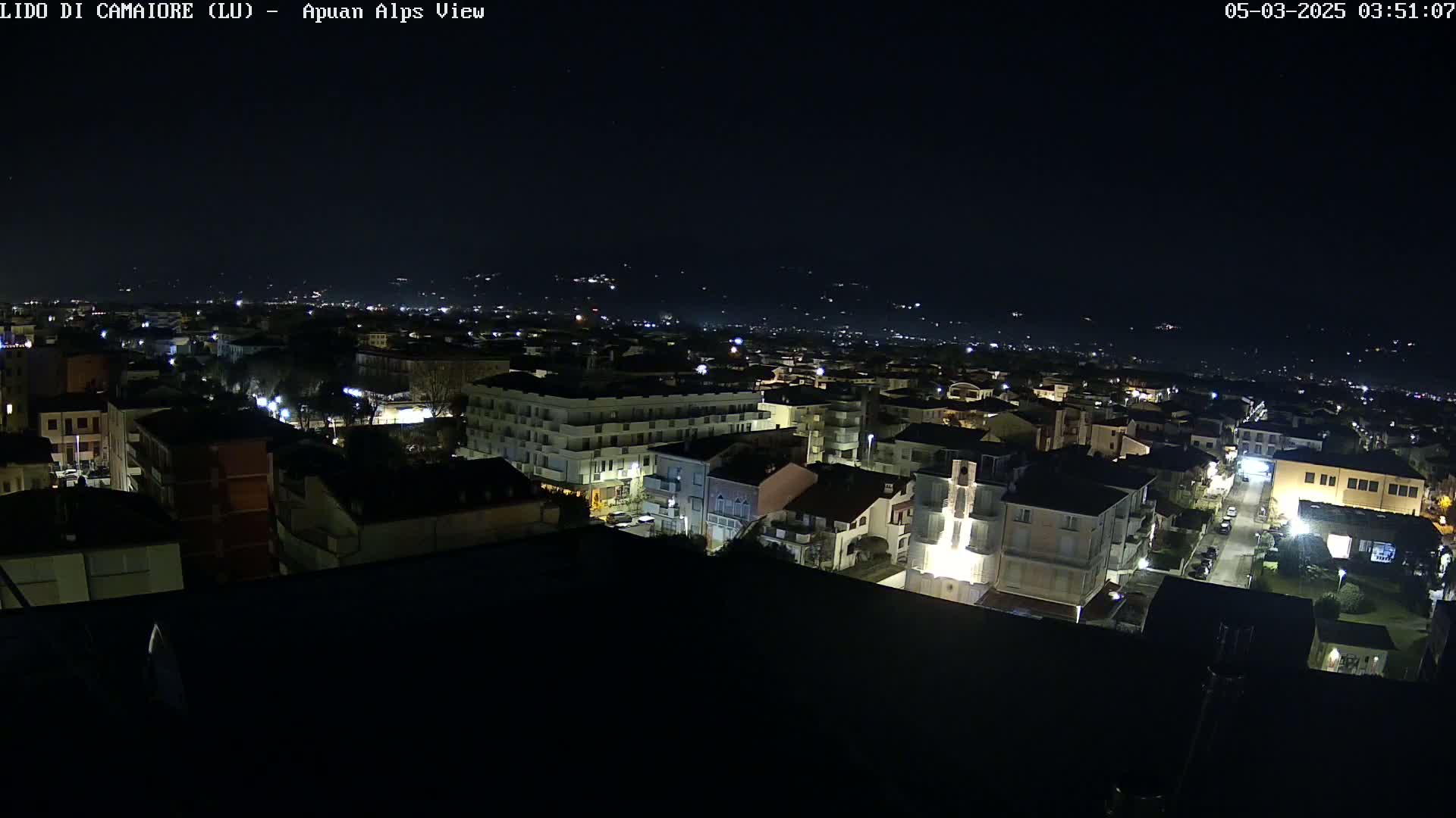 A nighttime aerial view of a town with illuminated buildings and streets, extending to a distant, dimly lit mountain range under a clear, dark sky.