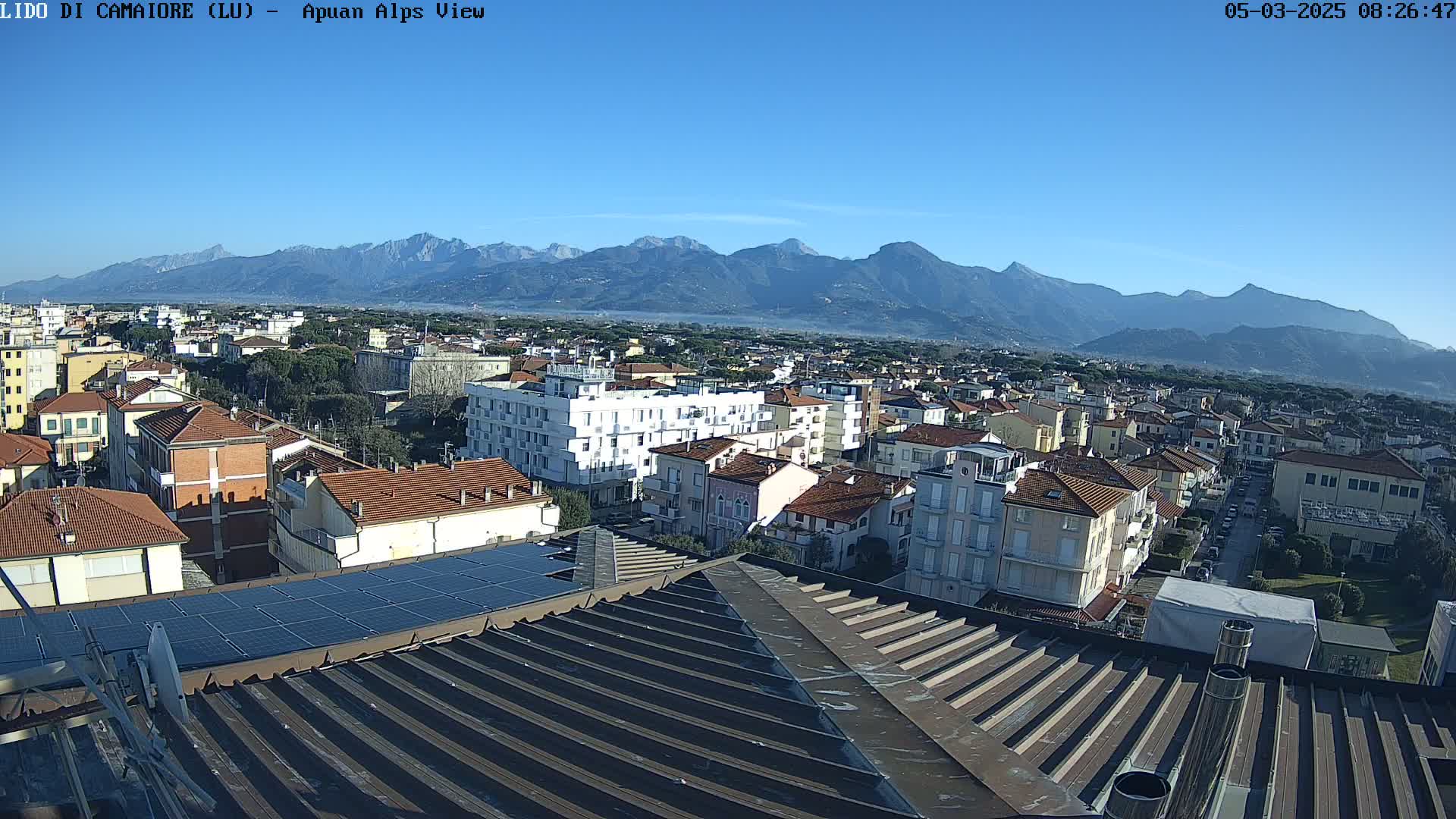 A clear, sunny day reveals a cityscape nestled at the foot of a mountain range, with solar panels visible on a rooftop in the foreground.