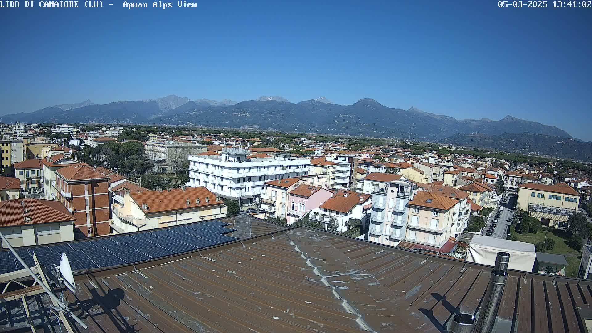 A rooftop view of a town with reddish-brown tile roofs, solar panels, and a clear blue sky above a mountain range in the distance.