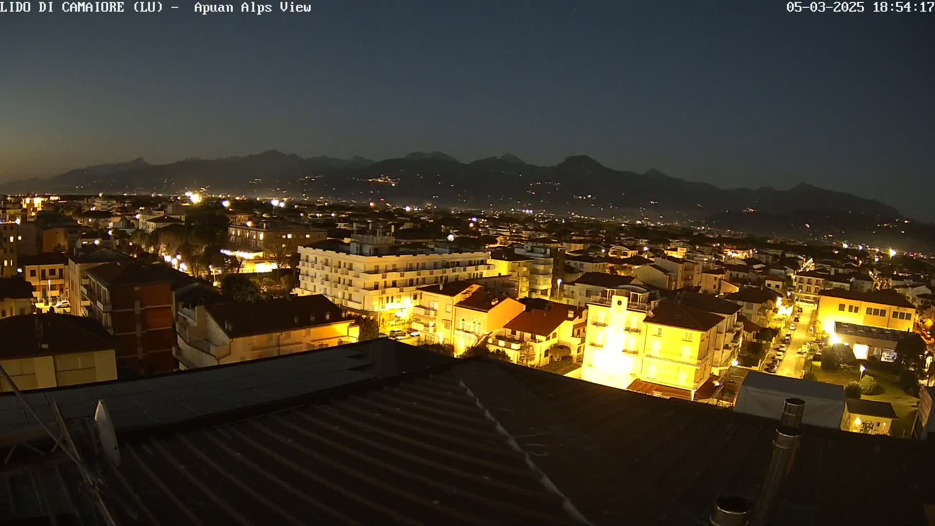 A nighttime view of a town illuminated by streetlights and building lights, with dark mountains in the background under a clear, dark sky.