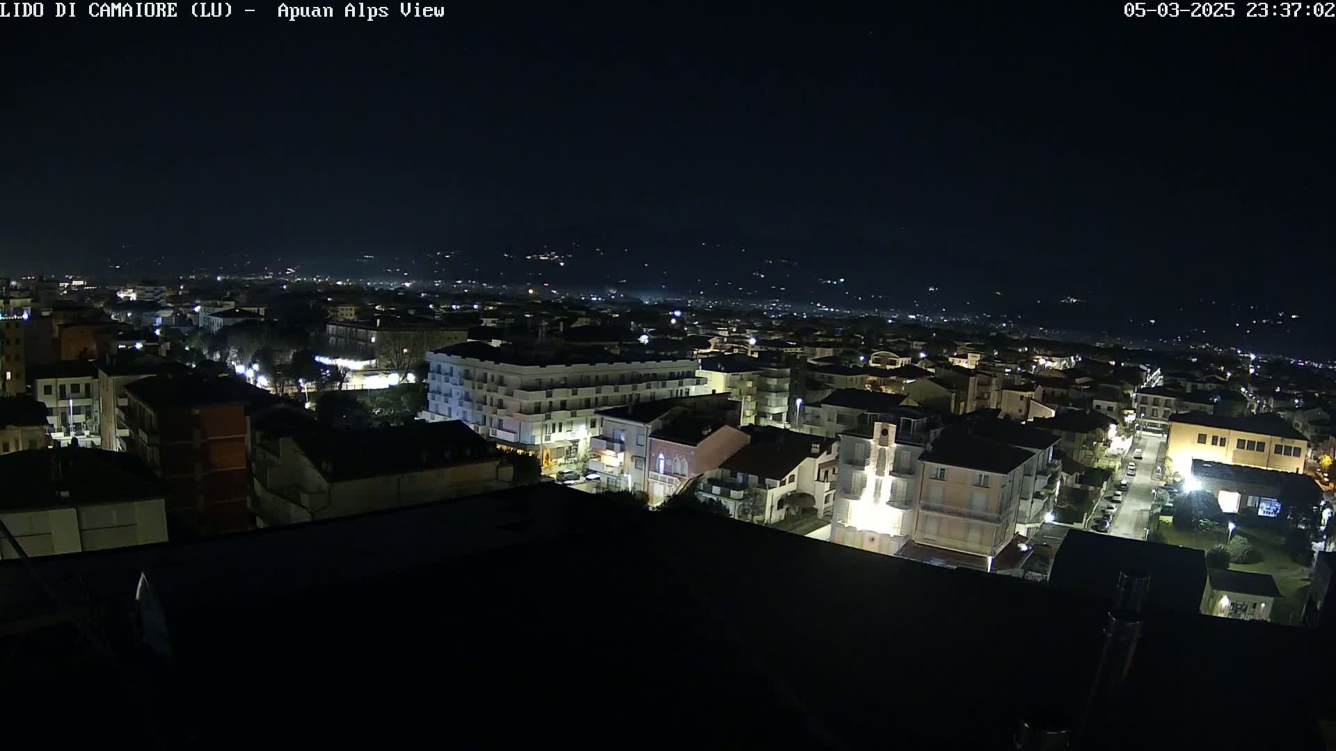A nighttime aerial view of a town illuminated by streetlights, with a dark sky and distant hills faintly visible in the background.