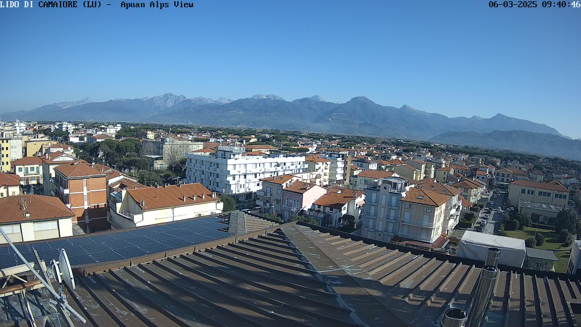 A clear, sunny day reveals a sprawling town nestled beneath a distant, snow-capped mountain range.