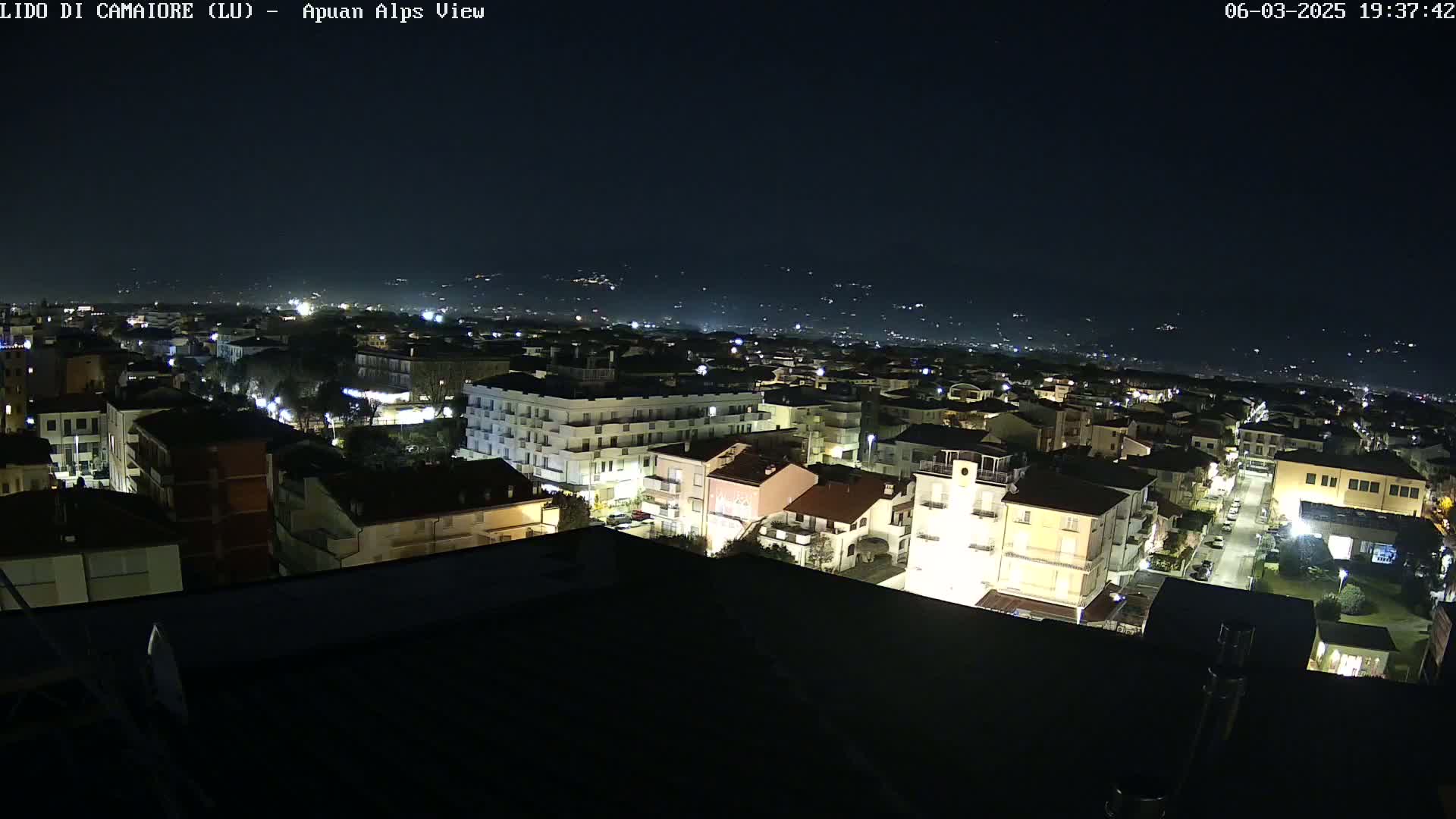 A nighttime aerial view of a town shows numerous buildings illuminated, with a clear, dark sky above.