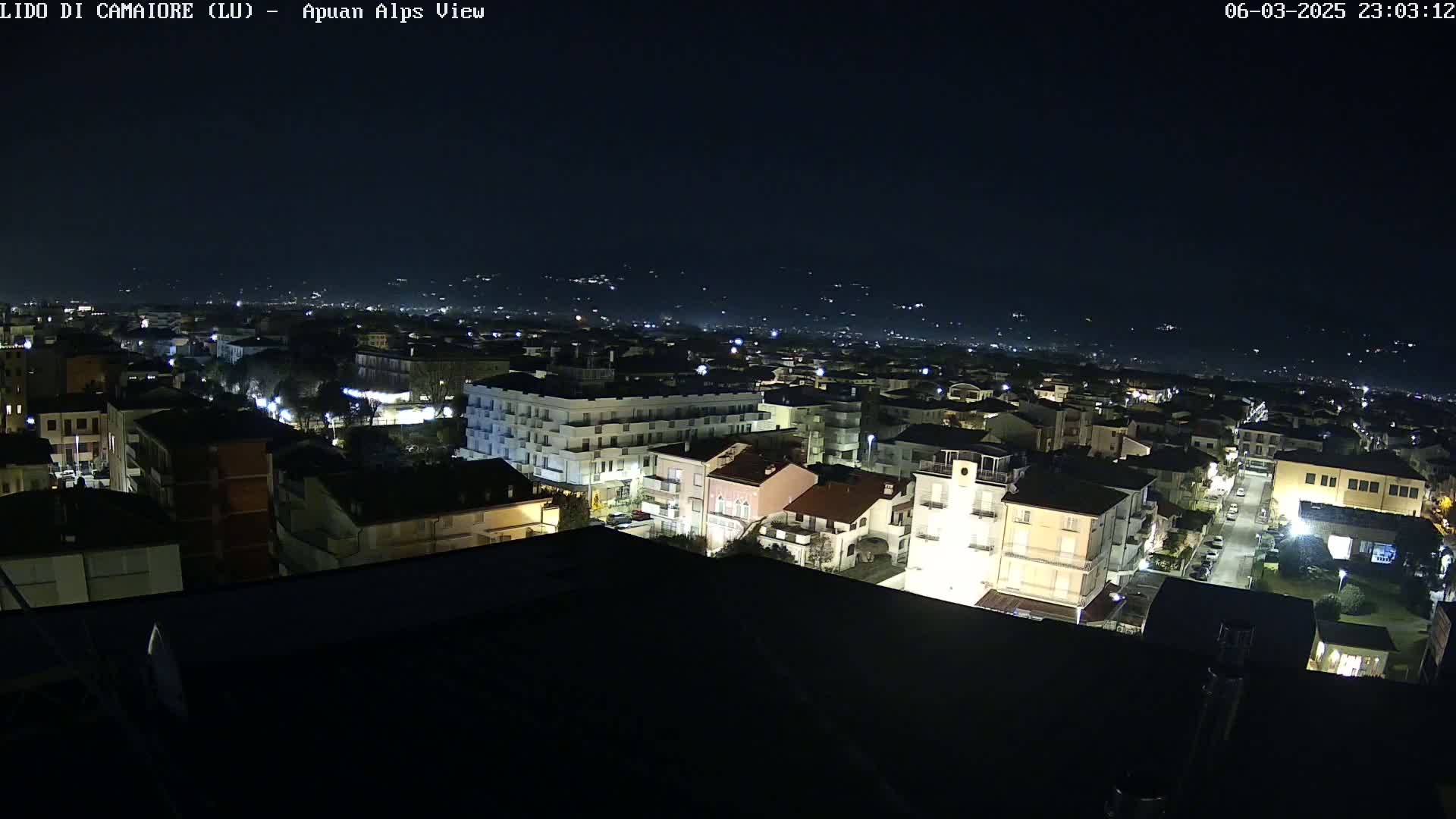 A nighttime aerial view of a town illuminated by streetlights, with distant hills faintly visible under a dark sky.