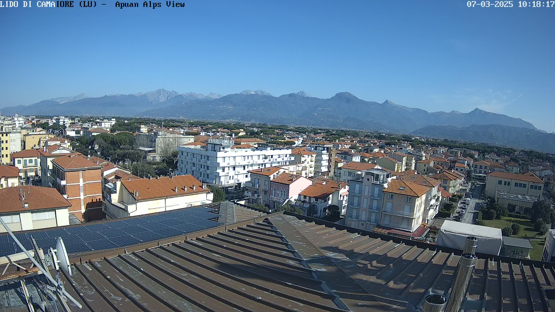 A clear, sunny day overlooks a town of low-rise buildings with red-tiled roofs, a view extending to snow-capped mountains in the distance.