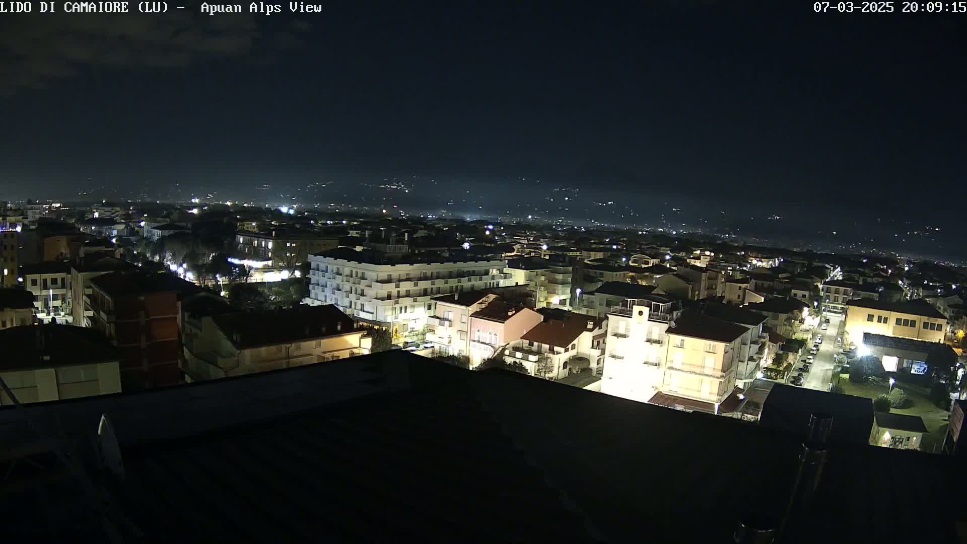 A nighttime aerial view of a town with illuminated buildings, streets, and distant hills under a clear, dark sky.