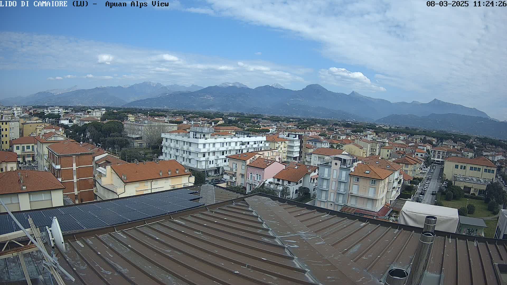 A partly cloudy day reveals a sprawling town nestled beneath a mountain range, viewed from a rooftop with solar panels.