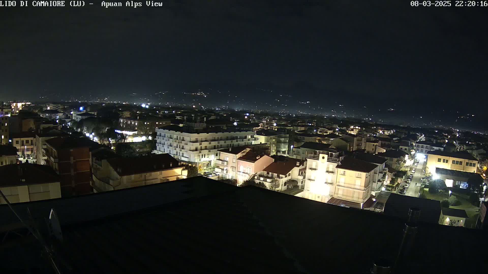 A nighttime, aerial view of a town, illuminated by streetlights and building lights, with a dark sky and distant hills faintly visible in the background.