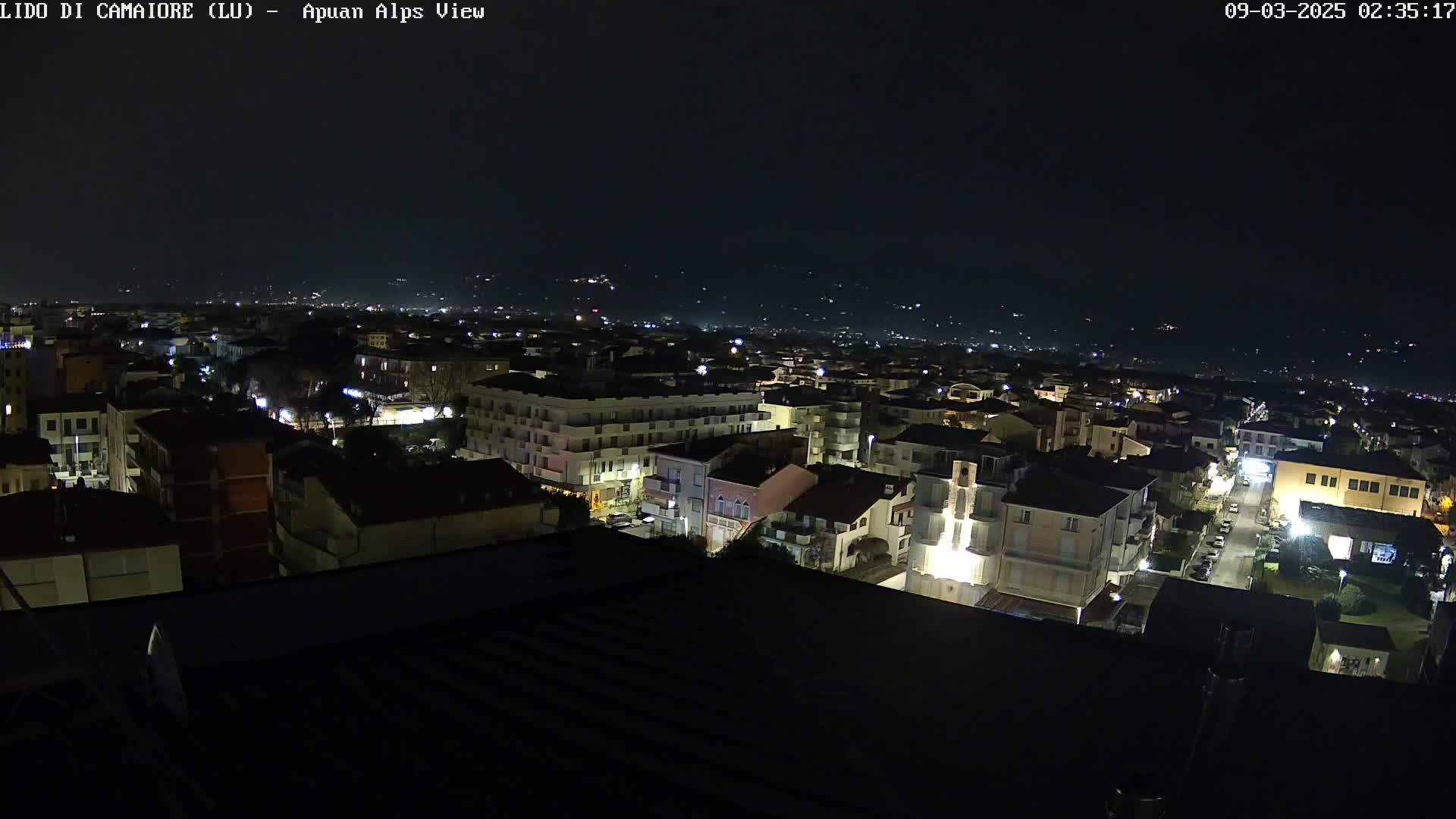 A nighttime aerial view of a town shows numerous buildings with illuminated windows, streets, and distant hills under a dark sky.