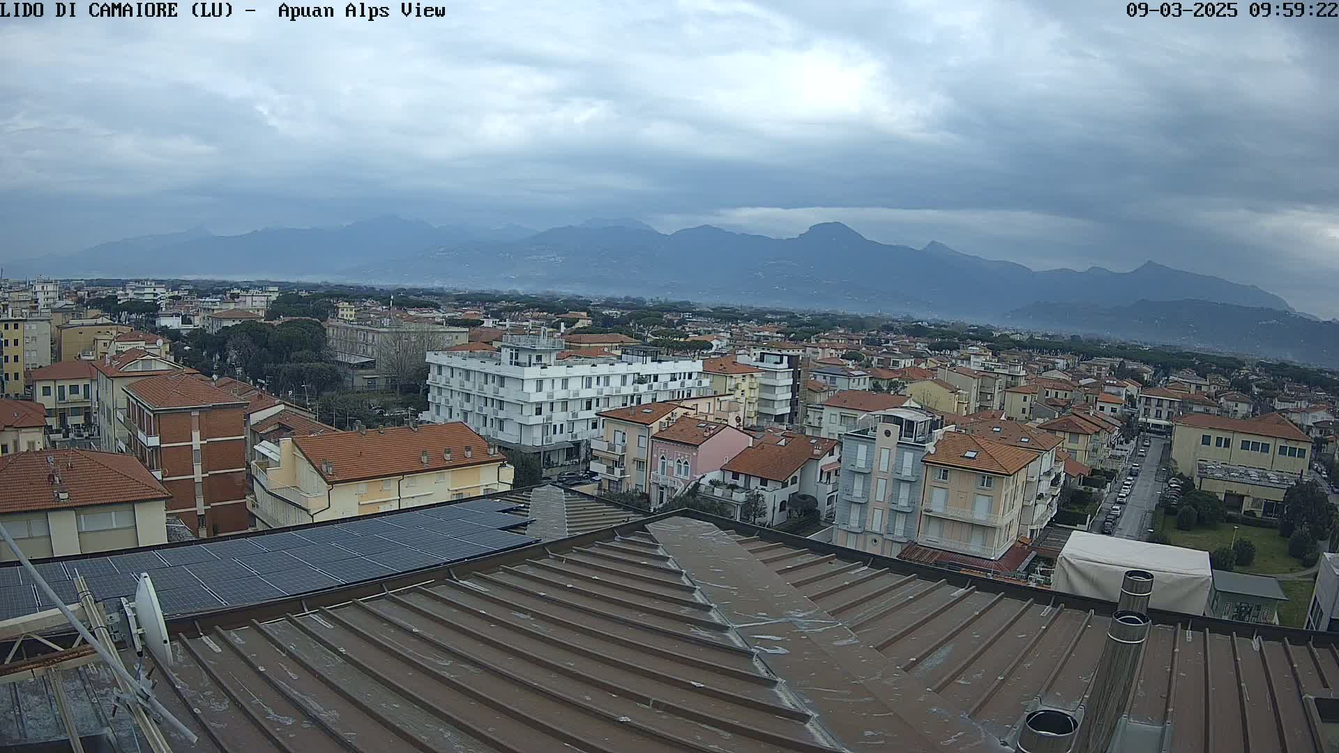 A view of a town nestled below a mountain range on an overcast day, seen from a rooftop with solar panels.