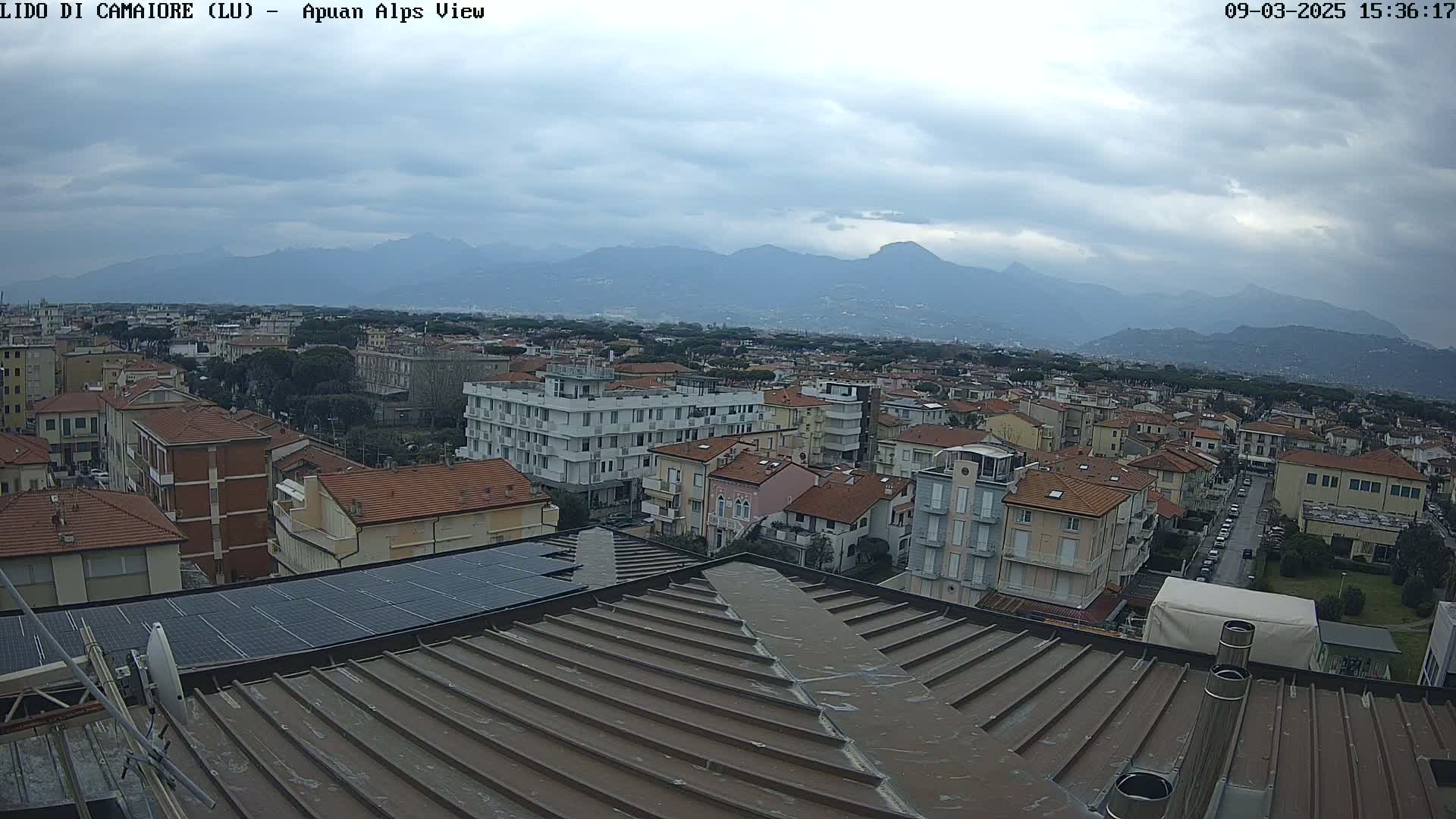 A view from a rooftop shows a town with many low-rise buildings under a cloudy sky, mountains in the background.