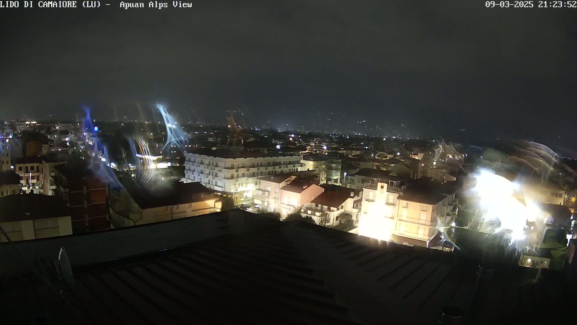 A nighttime, high-angle view of a town illuminated by streetlights, with streaks of light in the sky possibly indicating rain or wind.