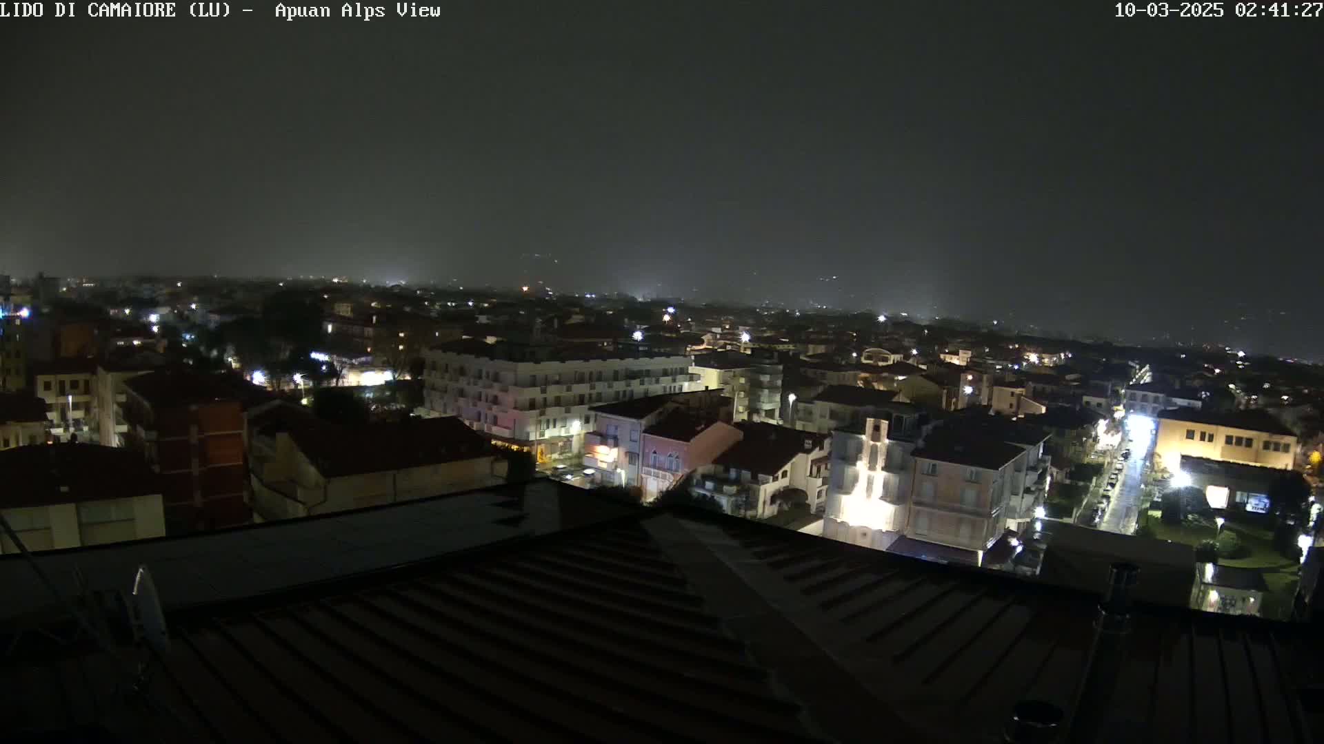 A nighttime aerial view of a town under a hazy, dark sky shows illuminated buildings and streets.